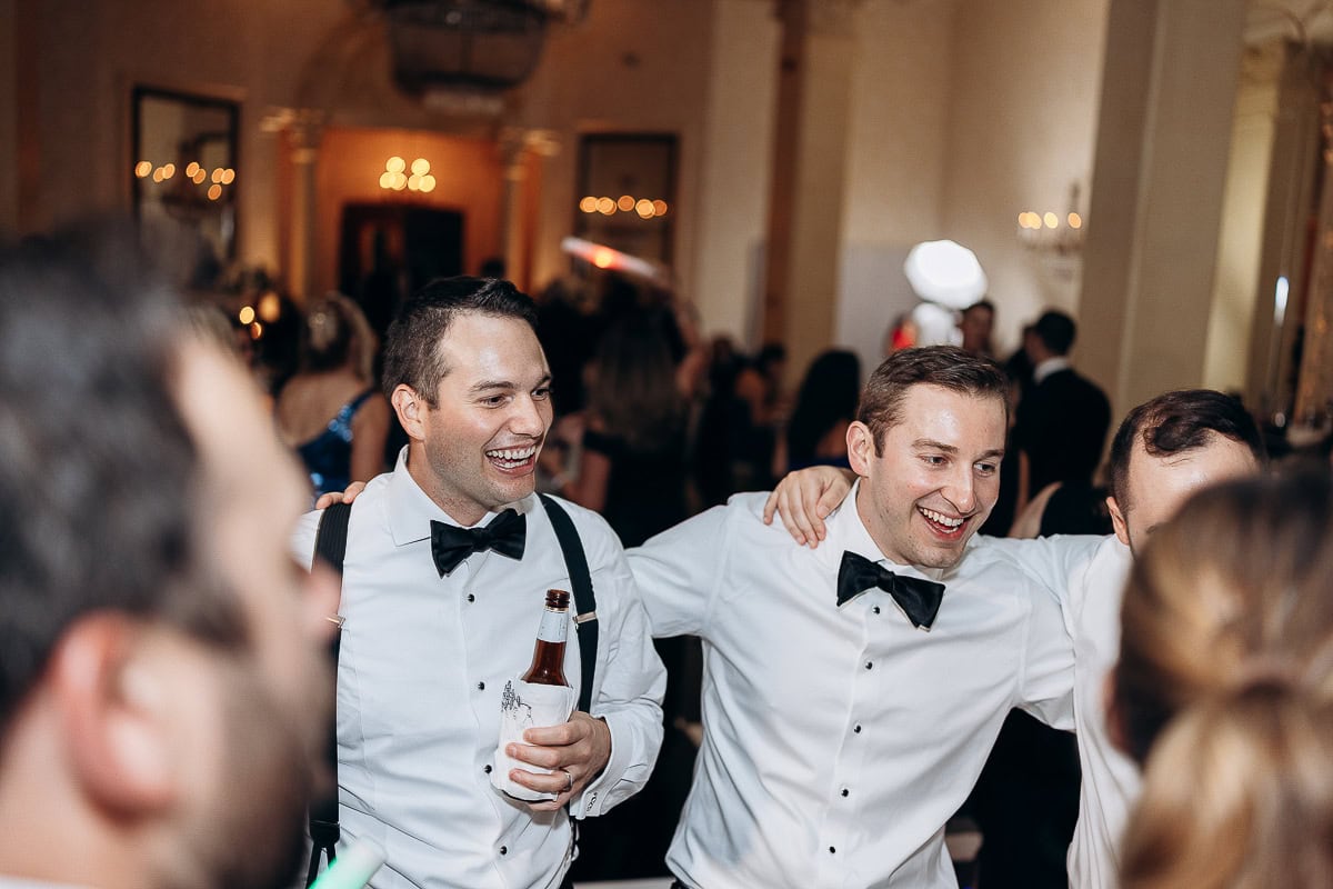 Two groomsmen in white shirts, black bow ties, and suspenders smiling and embracing during a wedding reception at Westchester Country Club, Rye, NY.