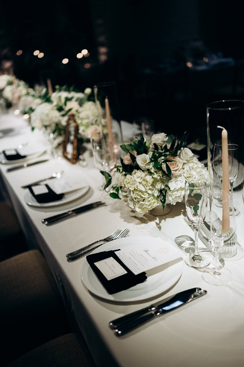 Elegant wedding table setting with white hydrangeas, blush roses, tapered candles in glass holders, white plates, and silver cutlery at Westchester Country Club.