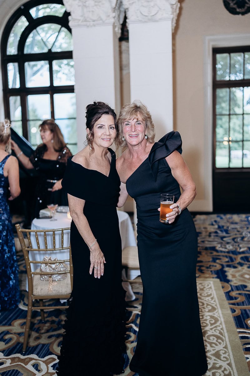 Two women in black evening gowns smiling and posing in a grand room with arched windows and patterned carpet at Westchester Country Club, Rye, NY.
