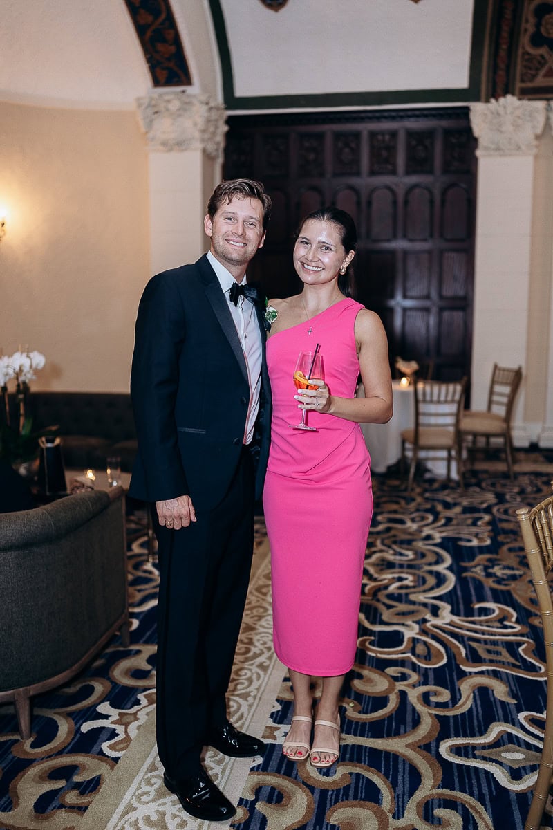 Man in black tuxedo and woman in pink one-shoulder dress standing on ornate carpet inside Westchester Country Club, Rye, NY, holding a cocktail glass.
