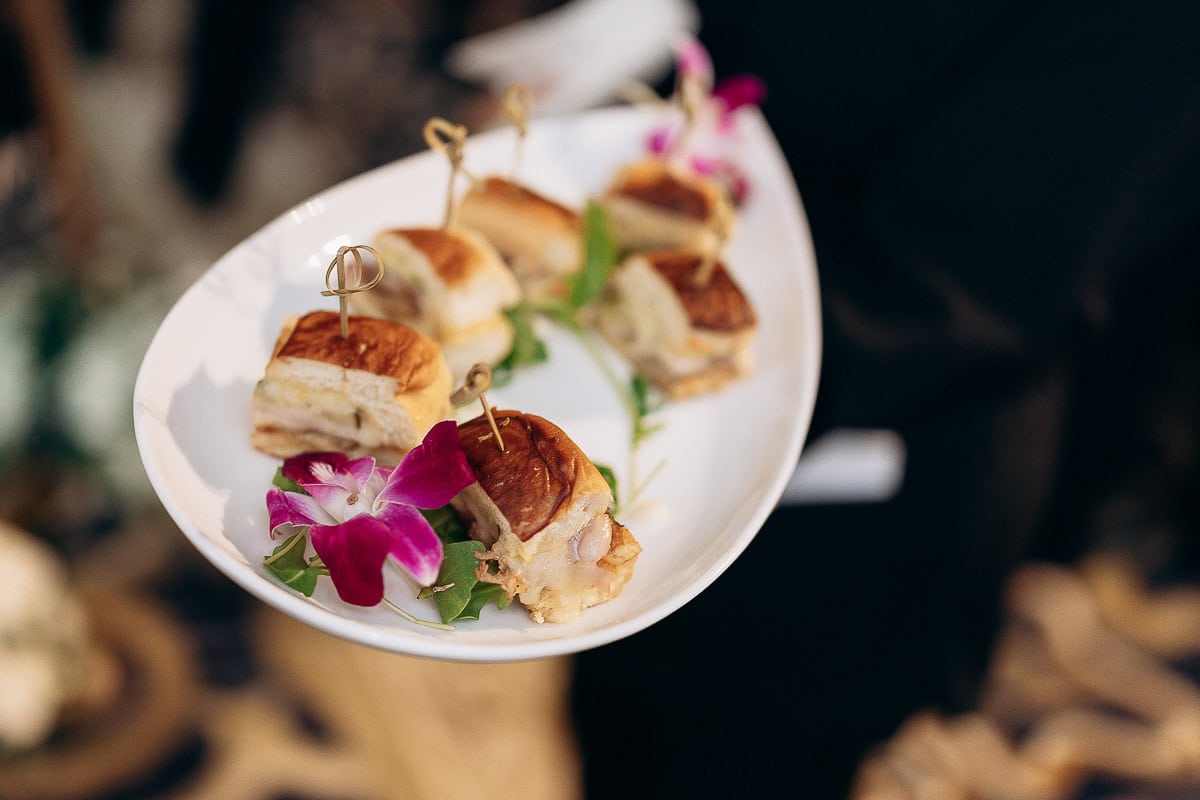 Close-up of a white platter holding toasted mini sandwiches with melted cheese, garnished with purple orchid flowers and green leaves, against a blurred background.