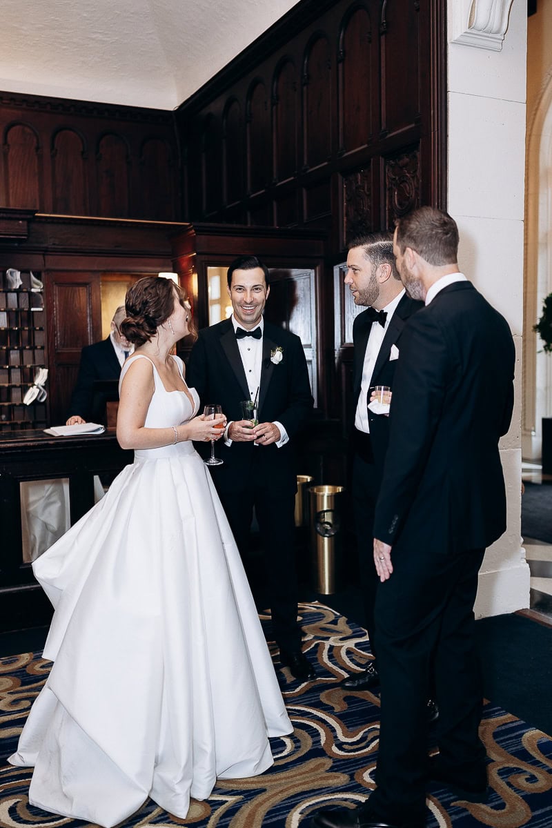 Bride in white gown and three men in black tuxedos conversing inside Westchester Country Club with dark wood paneling and patterned carpet.