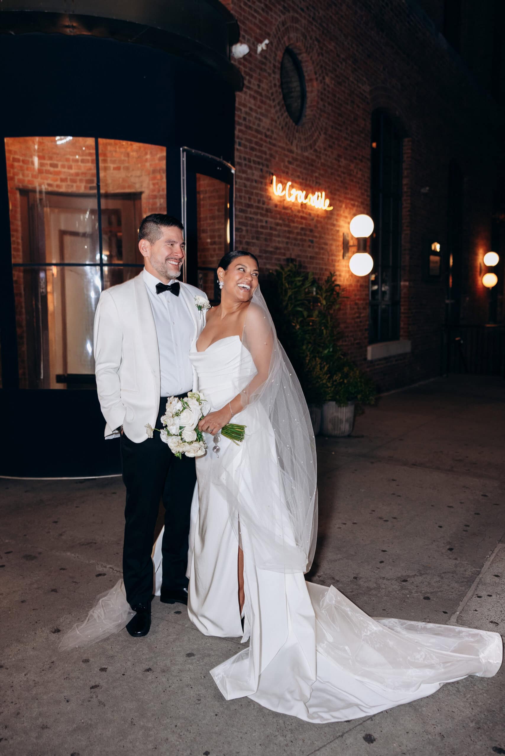 Bride and groom in elegant wedding attire standing outside Le Crocodile restaurant at night, brick wall and glowing street lamps in background.