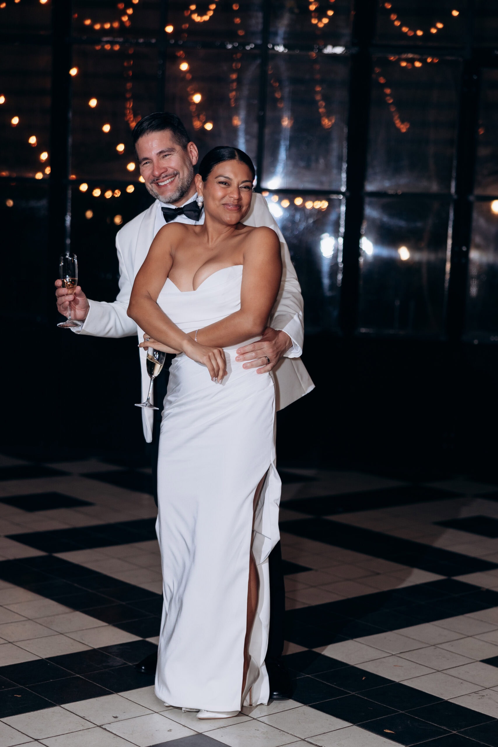 Couple in elegant white attire sharing an intimate embrace while holding champagne glasses, with warm string lights in the background at Wythe Hotel, Brooklyn.