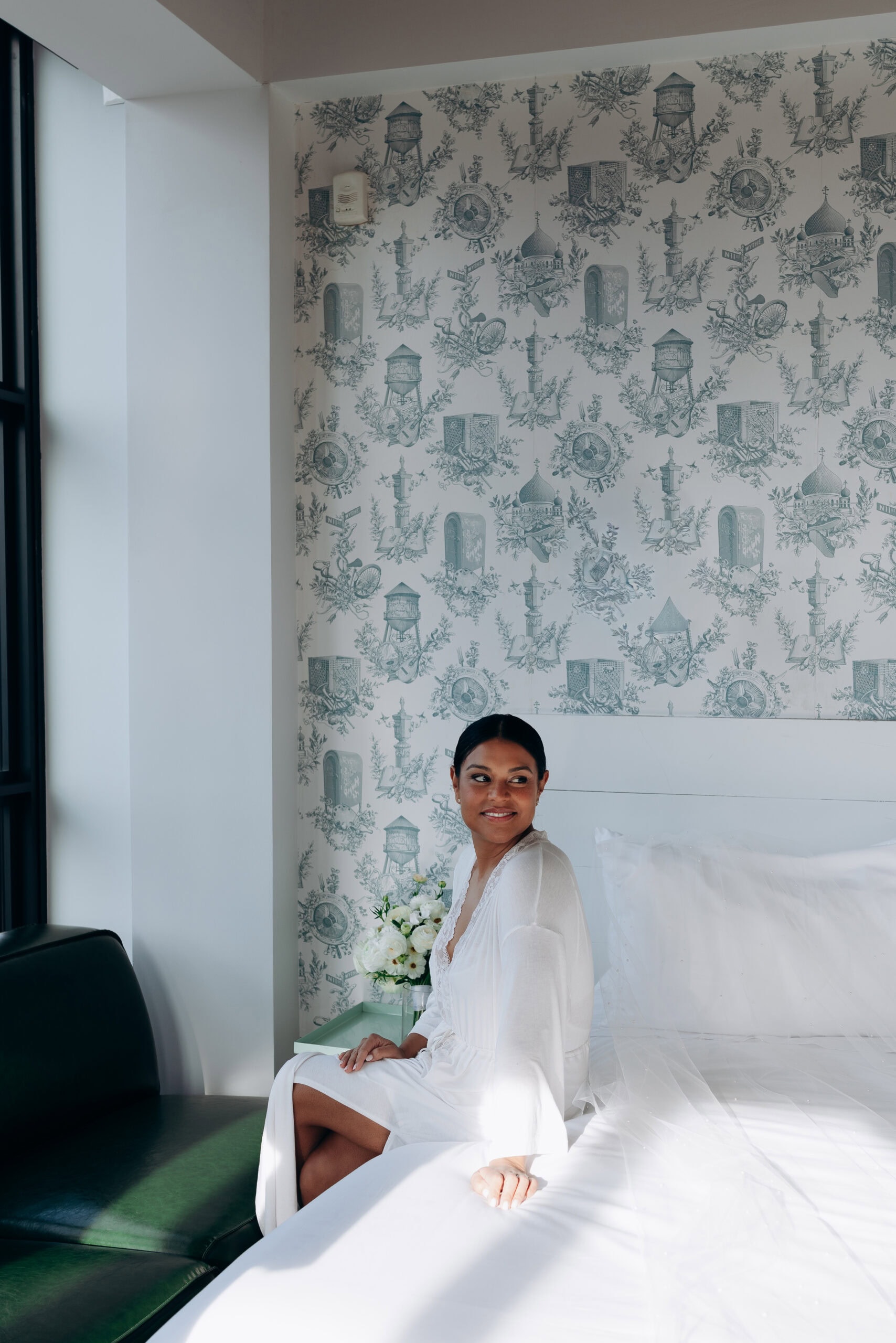 Bride in white robe sitting on bed with veil and bouquet, detailed toile wallpaper background, natural light, Wythe Hotel Brooklyn