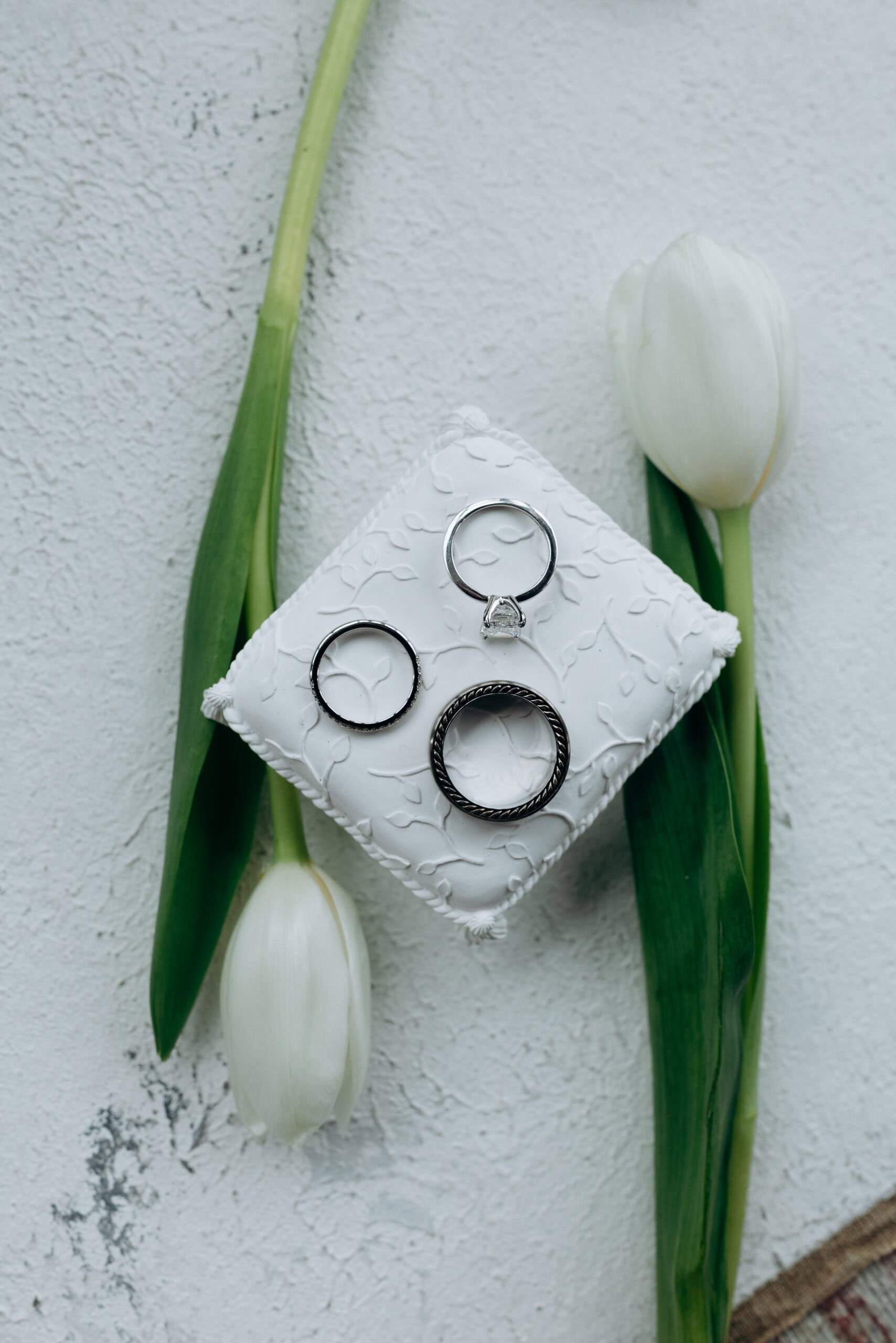Three silver wedding rings displayed on an embossed white cushion surrounded by two white tulips with green stems on a textured white surface.