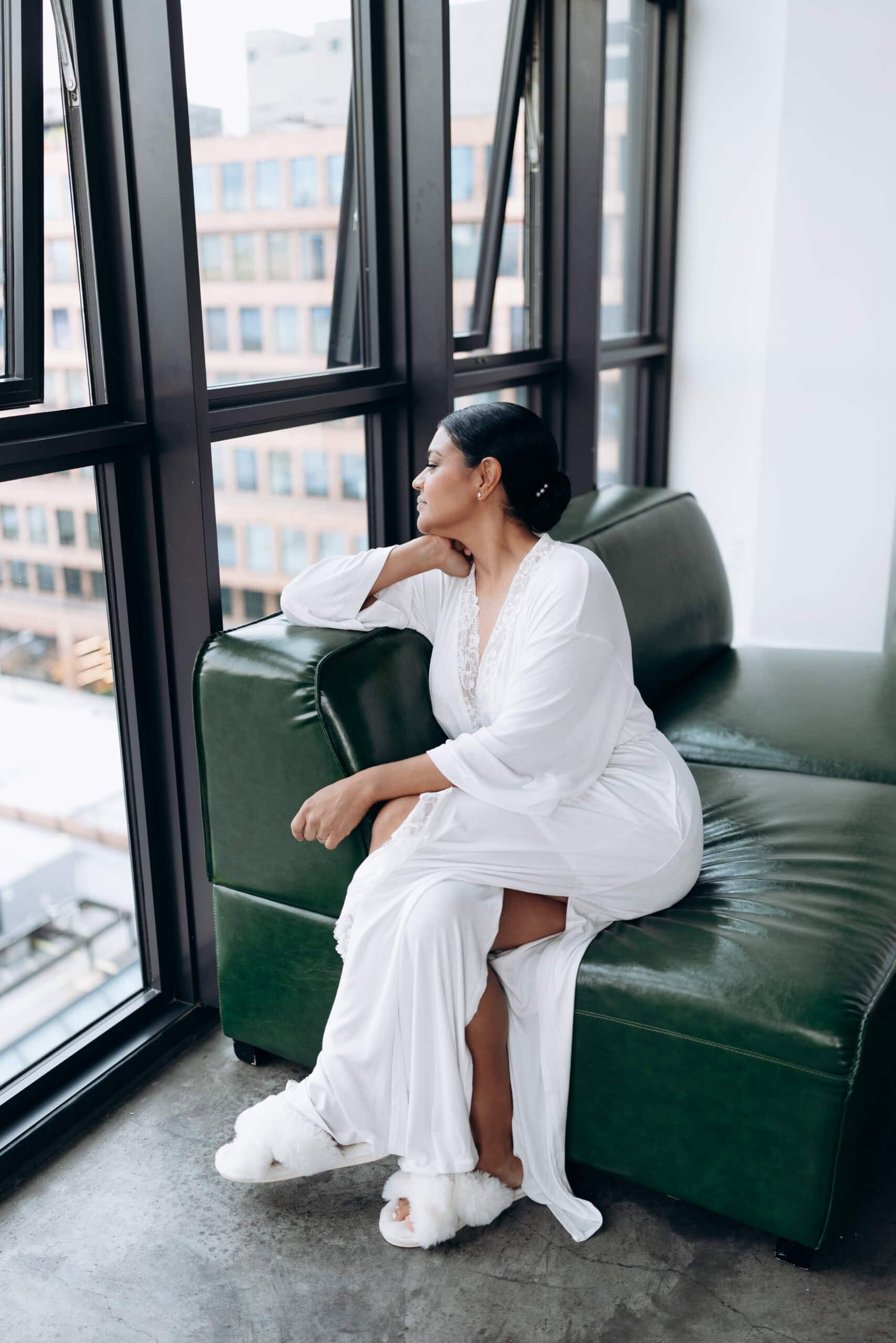 Woman in white lace-trimmed robe sitting on green leather sofa, gazing out large industrial windows at urban buildings in Brooklyn.