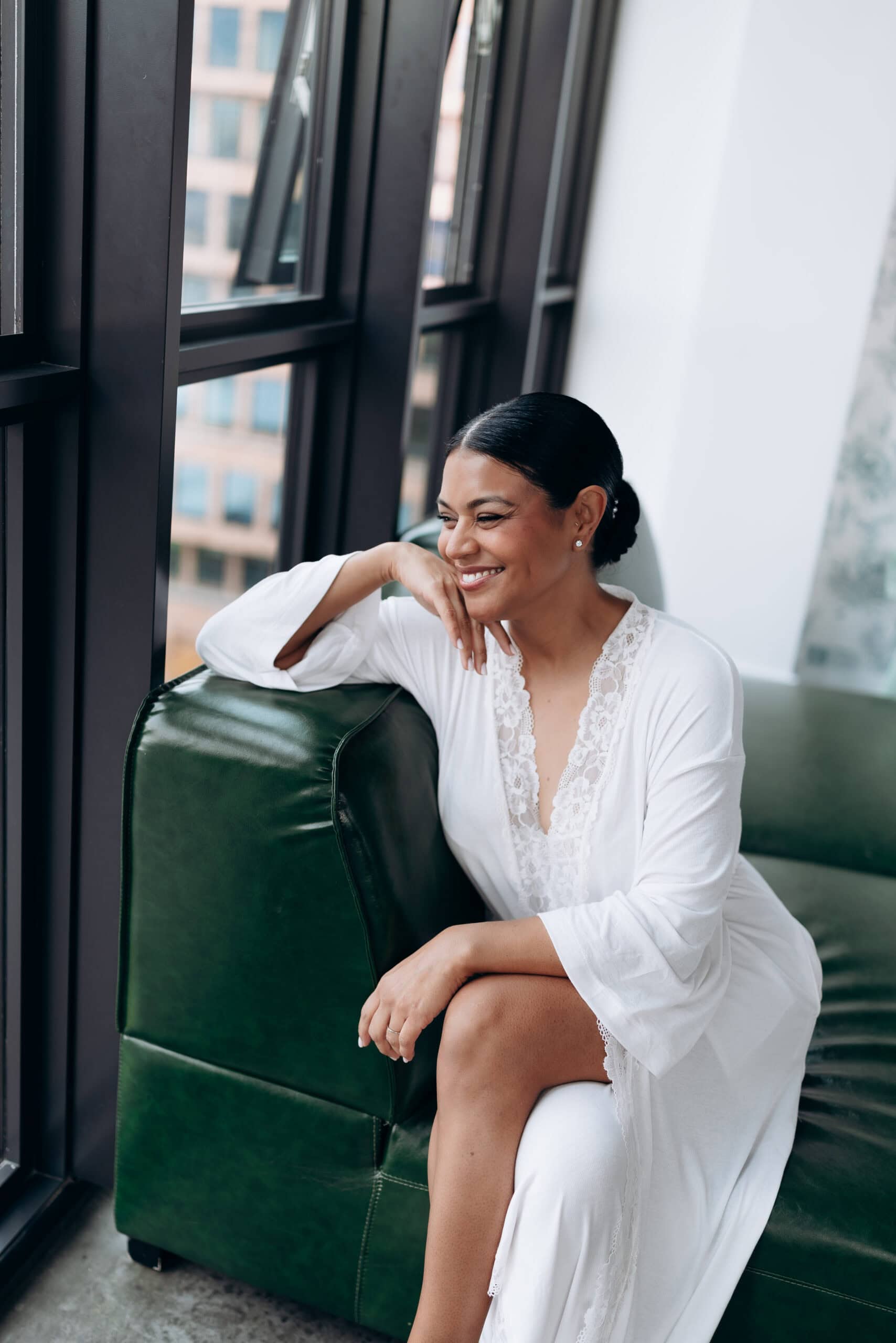 Woman in lace-trimmed white robe sitting on green leather sofa by large black-framed windows with city view at Wythe Hotel, Brooklyn.