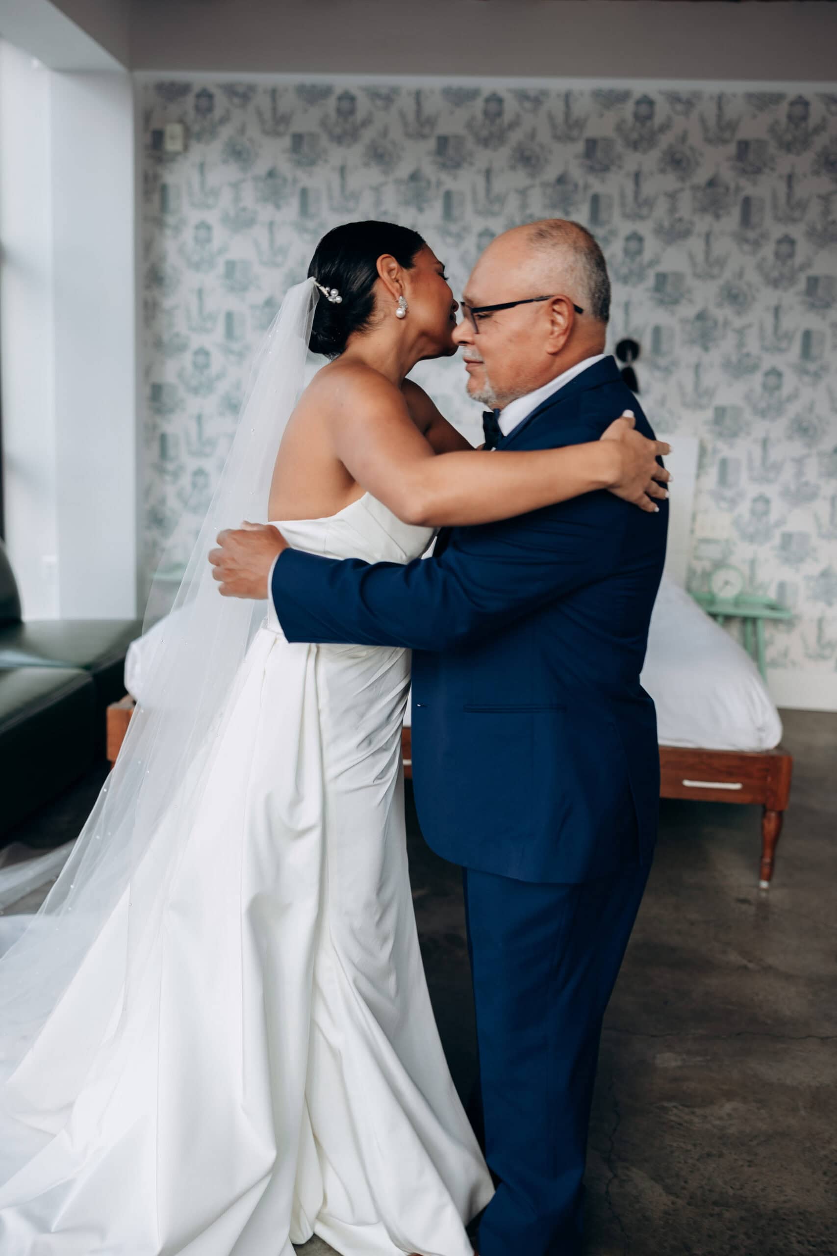 Bride in white satin wedding dress with veil embraces her father in a navy suit inside a softly lit room at Wythe Hotel, Brooklyn.