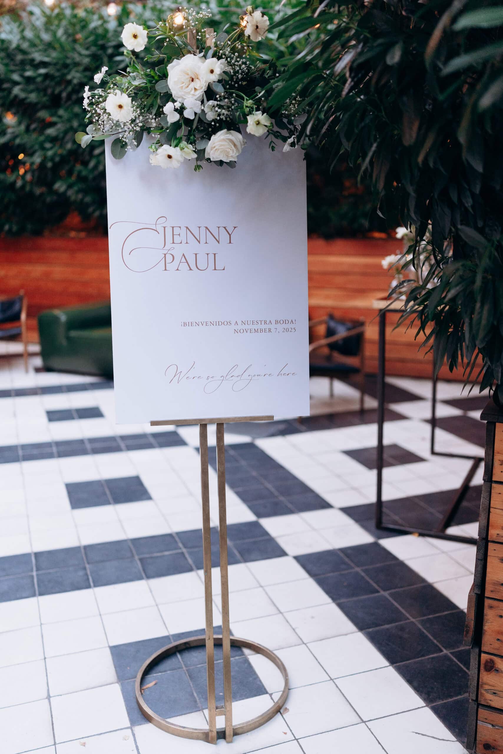 Wedding welcome sign featuring names Jenny and Paul with white floral arrangement on top, placed on a metal stand on black-and-white tiled floor with wooden wall and greenery in background.