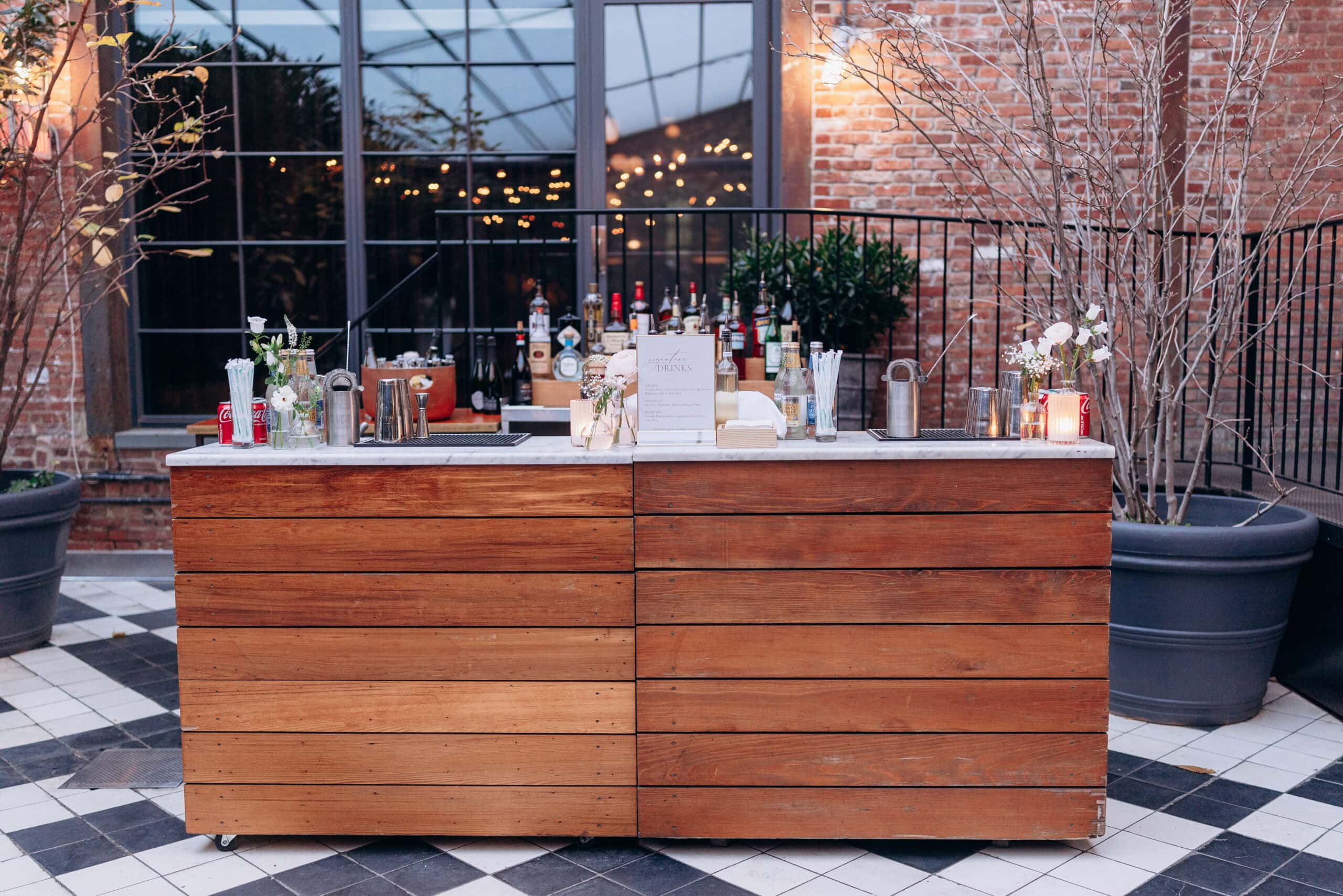 Outdoor wooden bar with marble top displaying assorted liquor bottles, glassware, and small floral arrangements, set against brick wall and large window at Wythe Hotel Brooklyn.