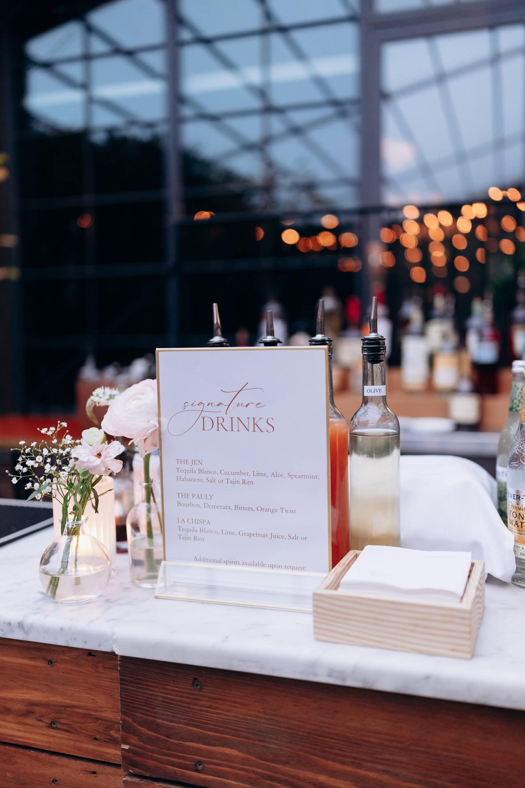 Marble bar counter with signature drinks menu, small floral vases, glass bottles, and warm bokeh lights in the background at an evening event.
