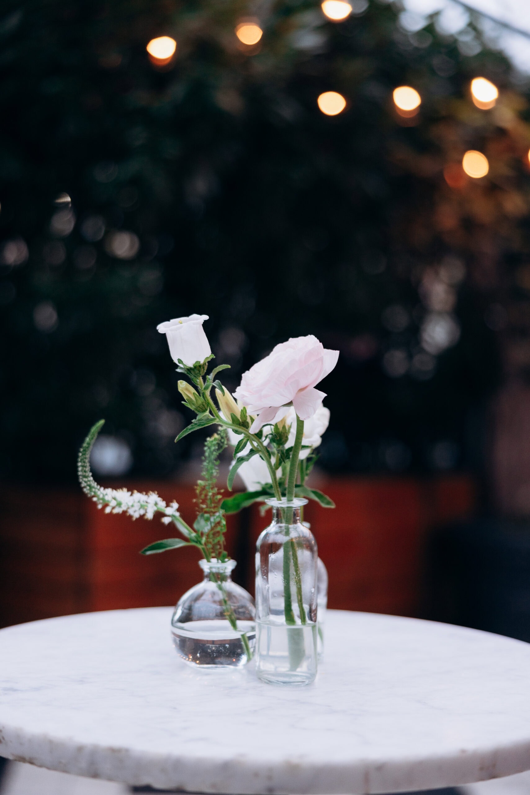Close-up of white flowers in clear glass vases on a marble table with blurred warm string lights in the background at Wythe Hotel, Brooklyn.