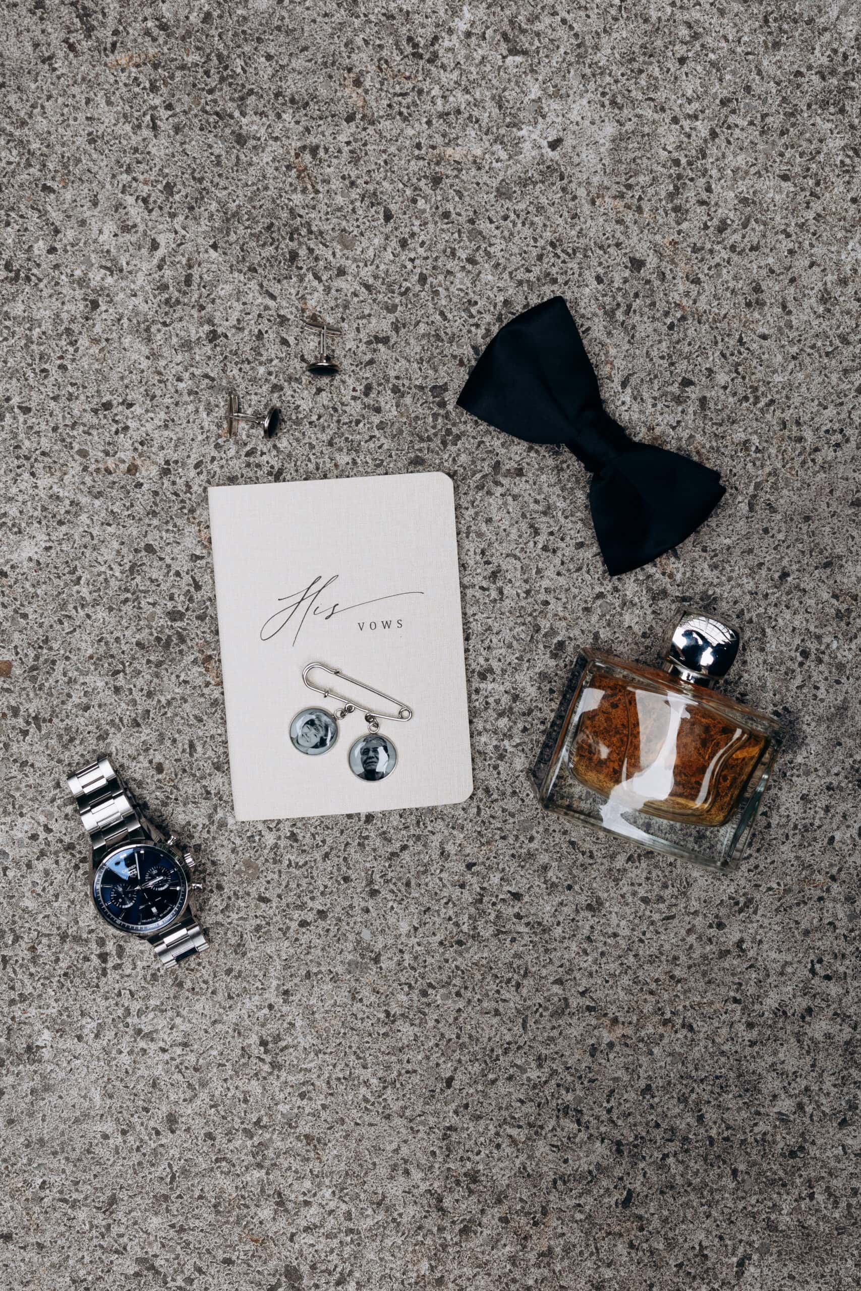 Flat lay of groom's accessories including black bow tie, 'His Vows' booklet with photo cufflinks, silver cufflinks, wristwatch, and cologne bottle on stone surface.