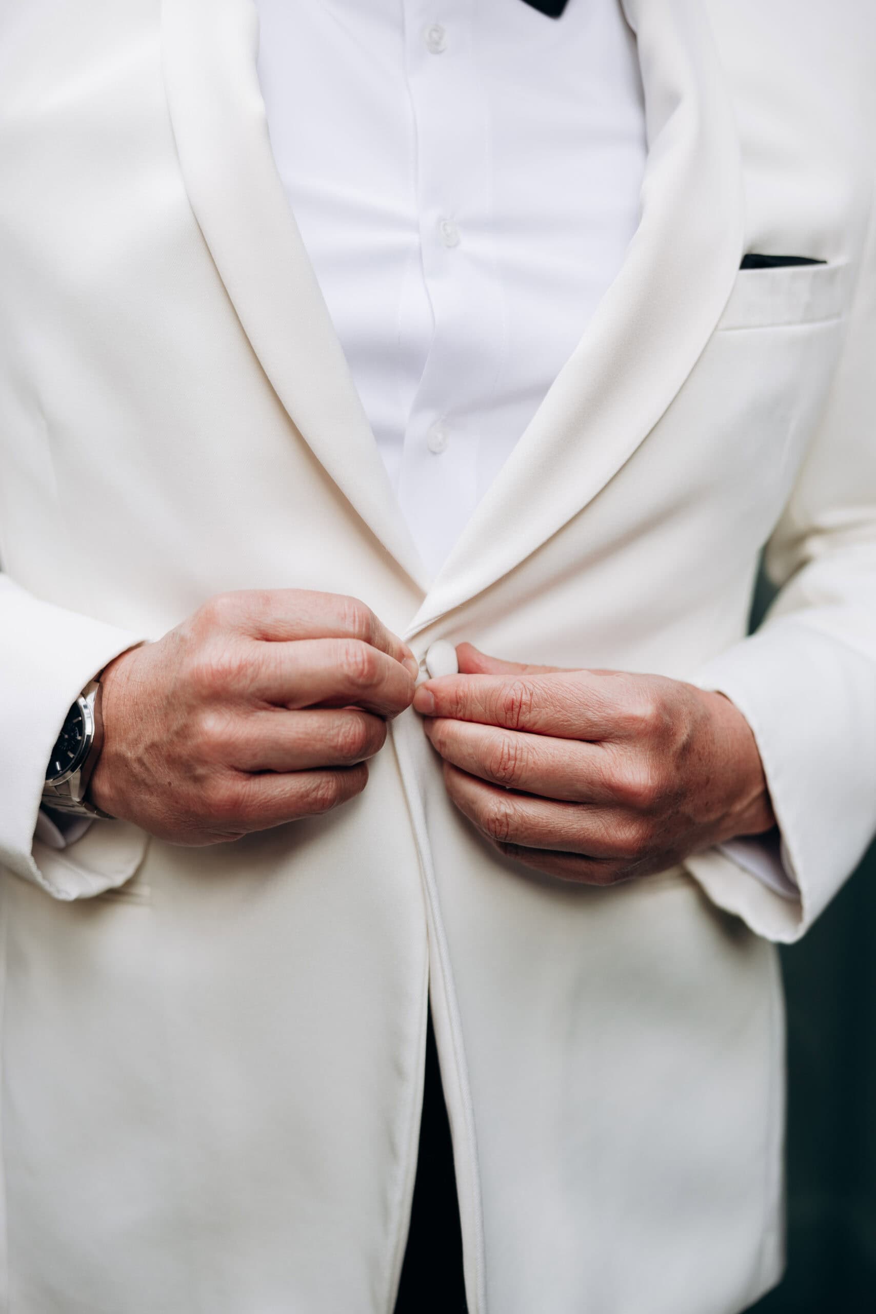 Close-up of a man's hands buttoning a white dinner jacket over a white dress shirt, emphasizing fabric texture and tailoring.