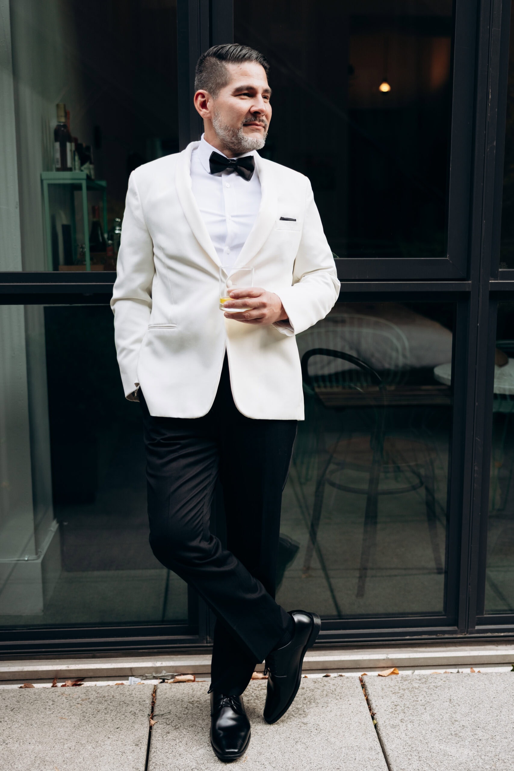 Man wearing a white dinner jacket, black bow tie, and black trousers, holding a glass of whiskey while leaning against a glass door outdoors.