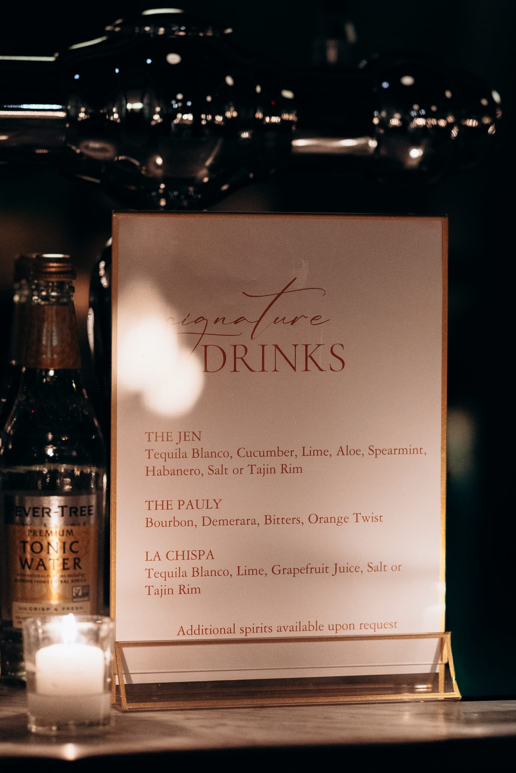 Close-up of a signature drinks menu on a bar counter with glass bottles, cocktail straws in a holder, and candlelight reflections in a dim setting at Wythe Hotel, Brooklyn.