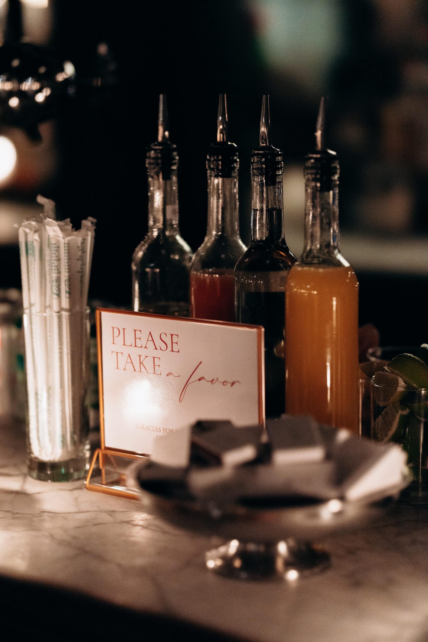 Close-up of glass bottles with pour spouts and a candlelit sign reading 'Please take a favor' on a marble bar surface in low warm lighting.