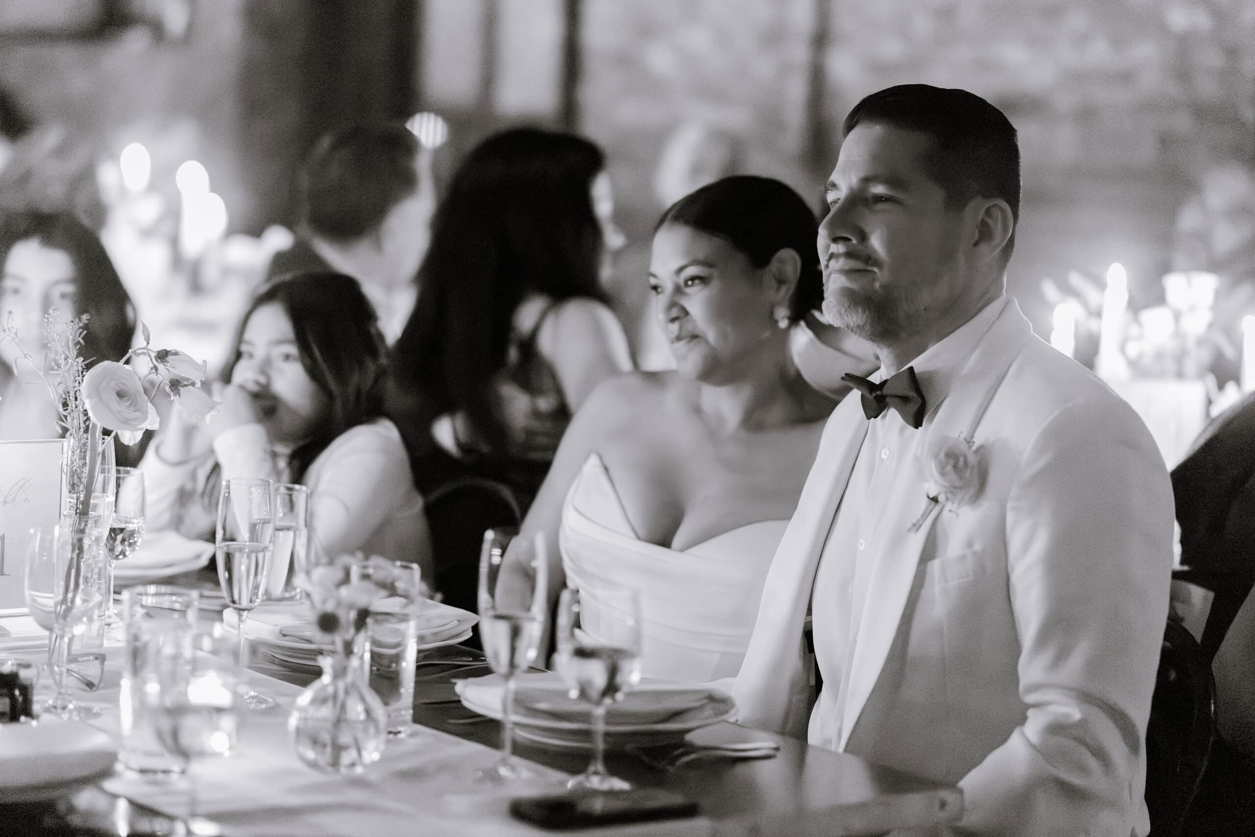 Bride and groom seated at a candlelit wedding reception table with floral centerpieces, guests blurred in the background at Wythe Hotel Brooklyn NY.