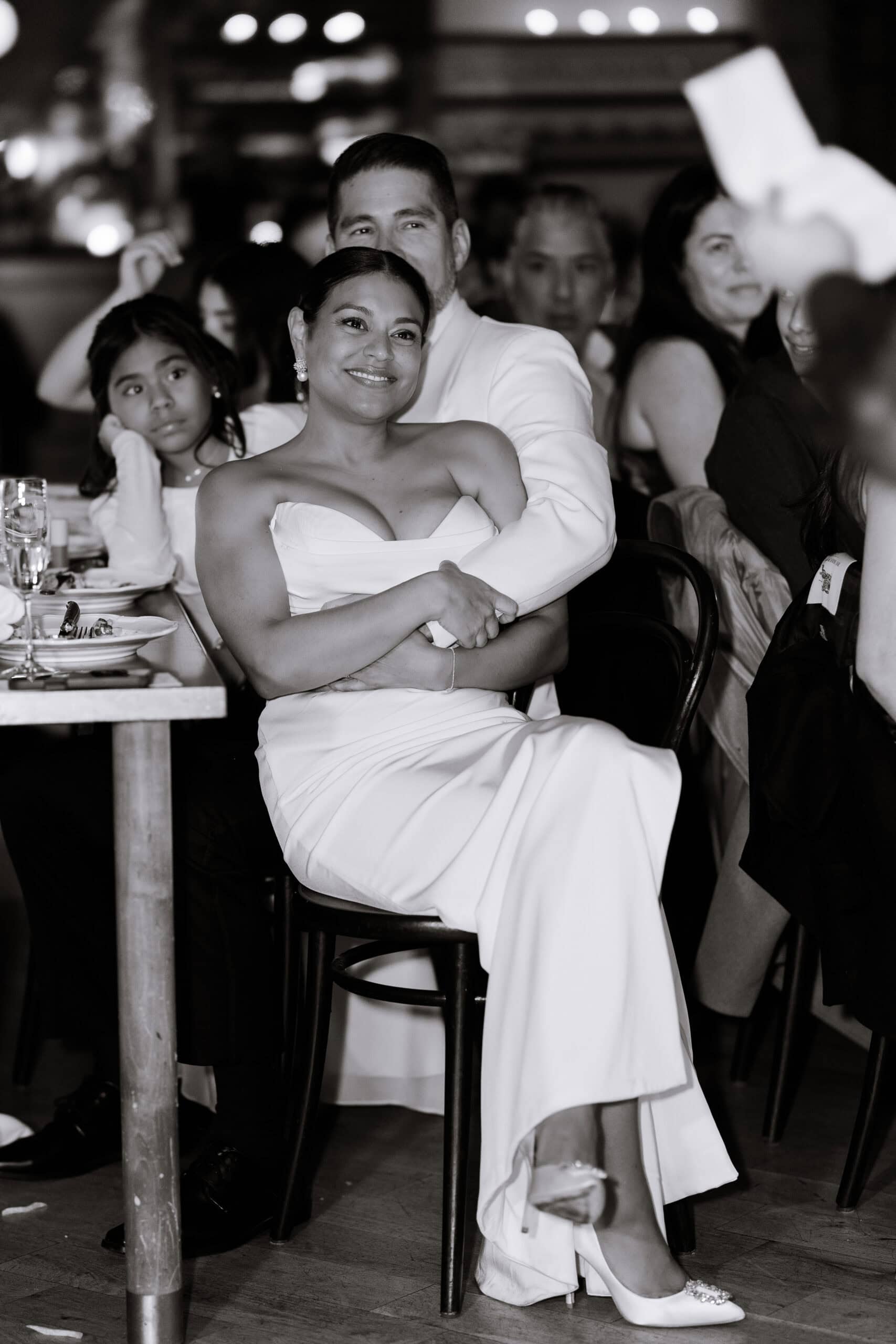Bride in white gown and embellished heels embraced by partner at a warmly lit wedding reception with guests seated nearby at Wythe Hotel, Brooklyn.