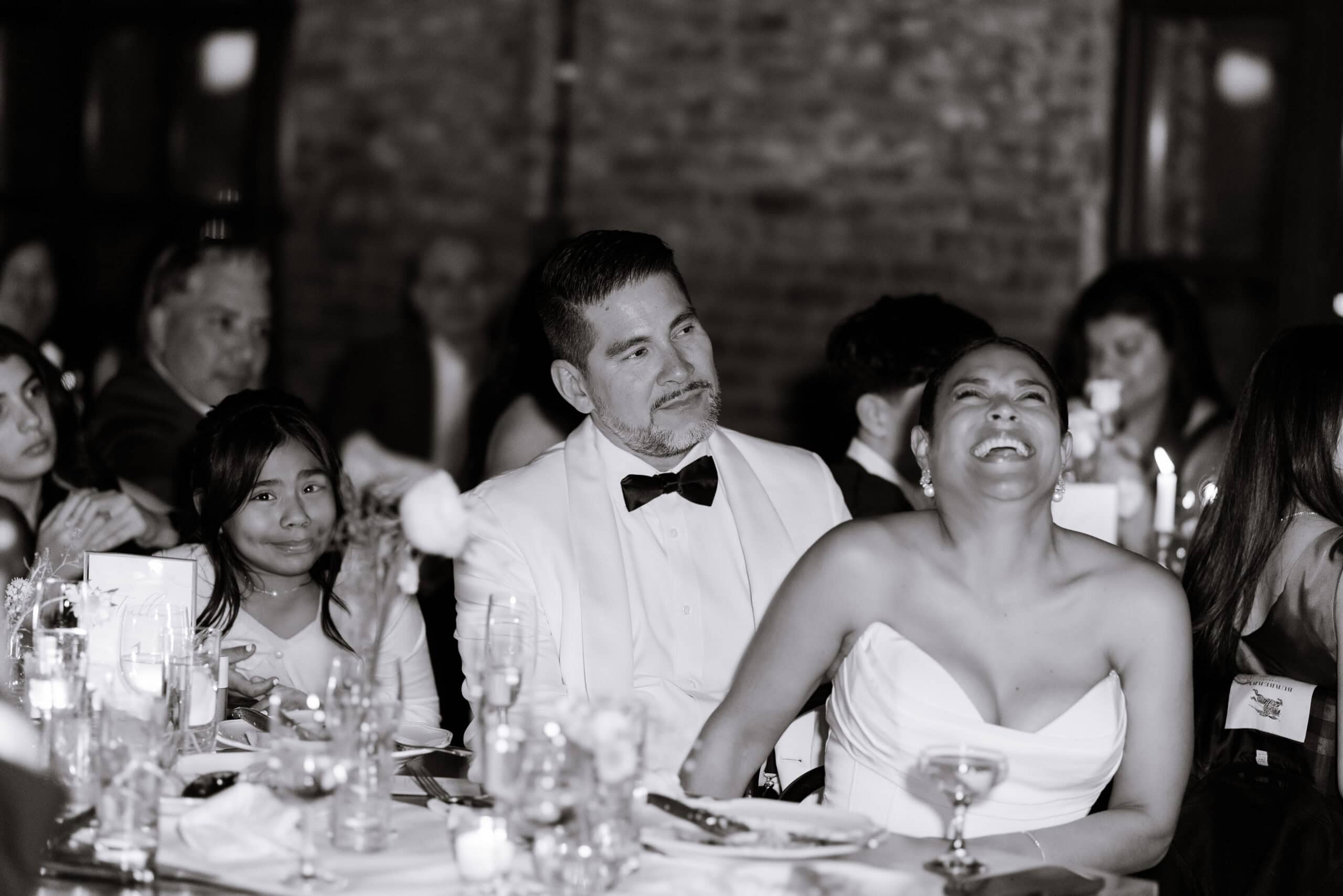Groom in white tuxedo looking at bride laughing joyfully during wedding reception at Wythe Hotel, Brooklyn, NY, surrounded by guests and candlelit table.