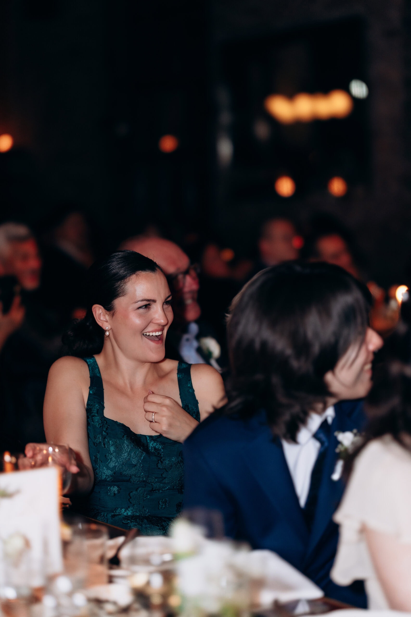 Woman in emerald lace dress laughing during a wedding reception at the Wythe Hotel, Brooklyn, NY, with softly lit background and guests.