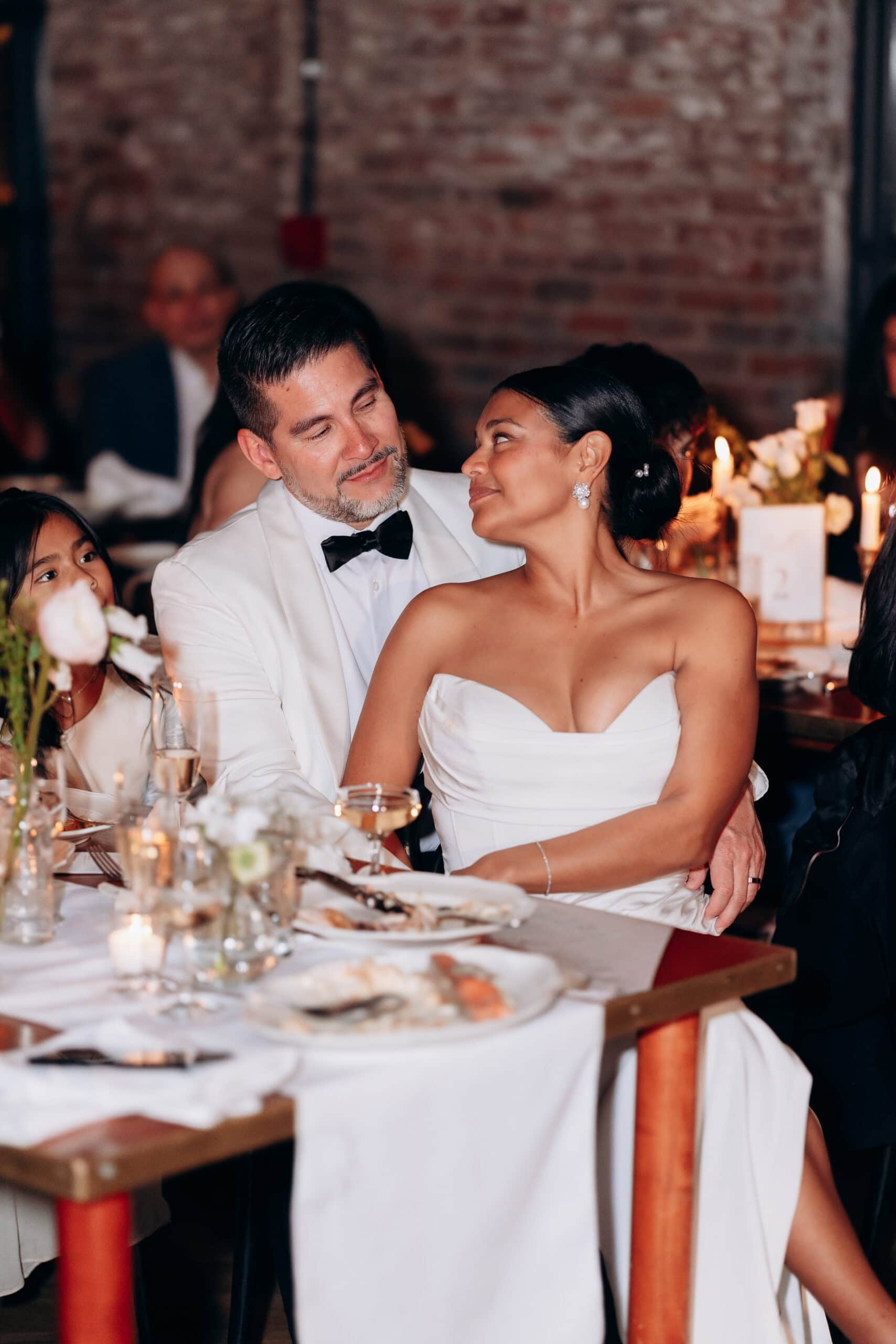 Bride and groom seated closely at a candlelit wedding reception table with white floral arrangements and exposed brick background at Wythe Hotel, Brooklyn.