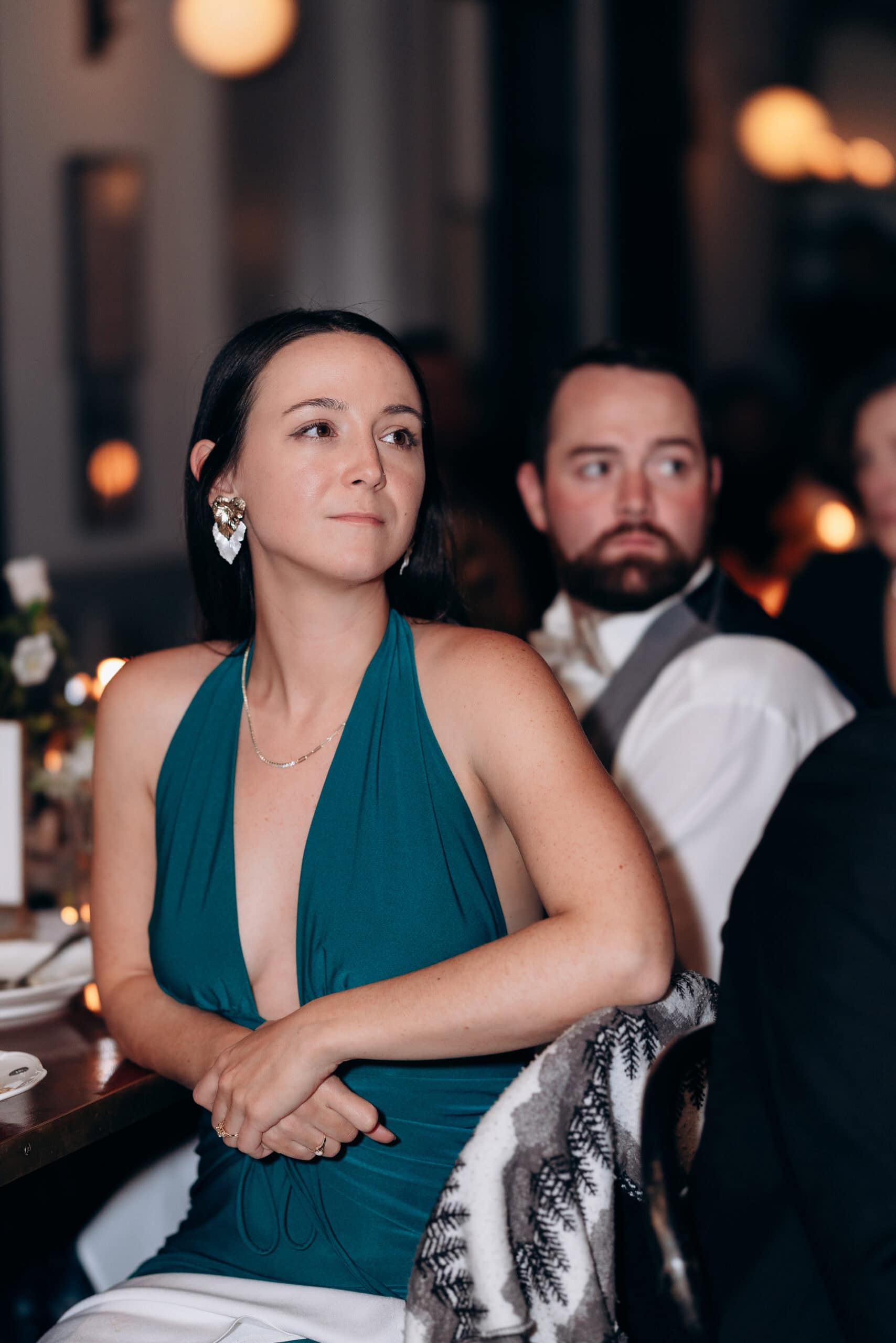Woman in teal dress and gold earrings seated at a table with a man blurred in the background, ambient warm lighting at Wythe Hotel, Brooklyn.