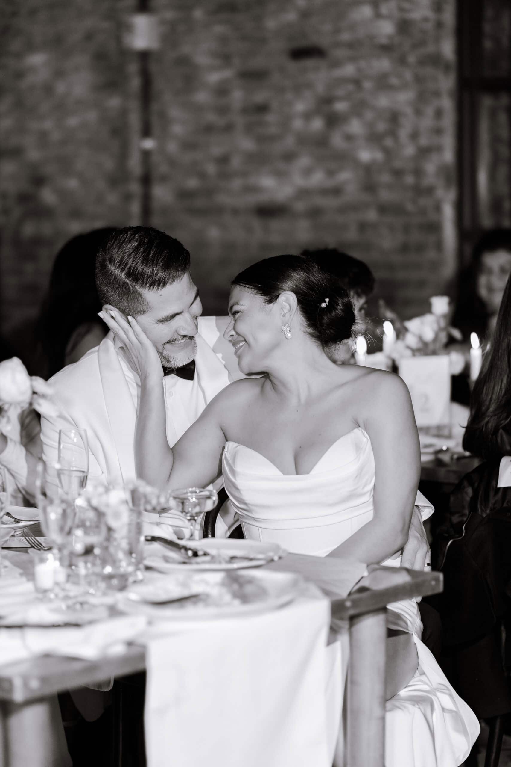 Bride and groom sharing an intimate smile at their wedding reception with a brick wall and candlelit table in the background at the Wythe Hotel, Brooklyn.