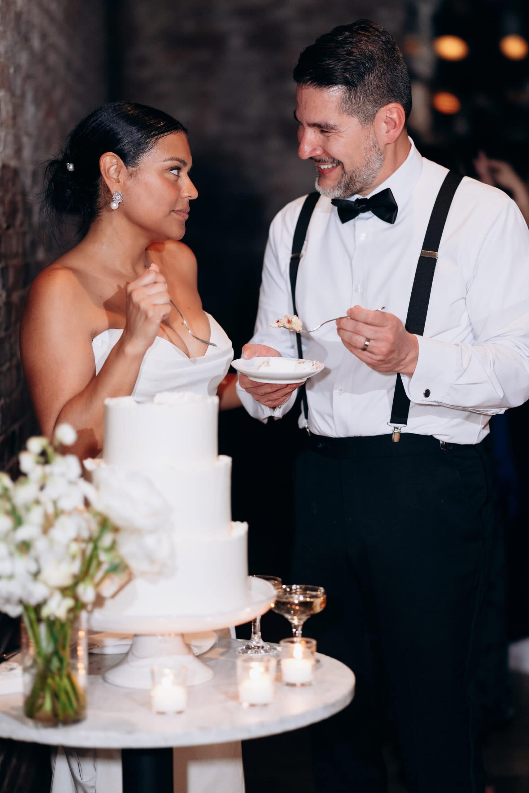 Bride and groom sharing a moment while feeding each other wedding cake near a white tiered cake and floral centerpiece at Wythe Hotel