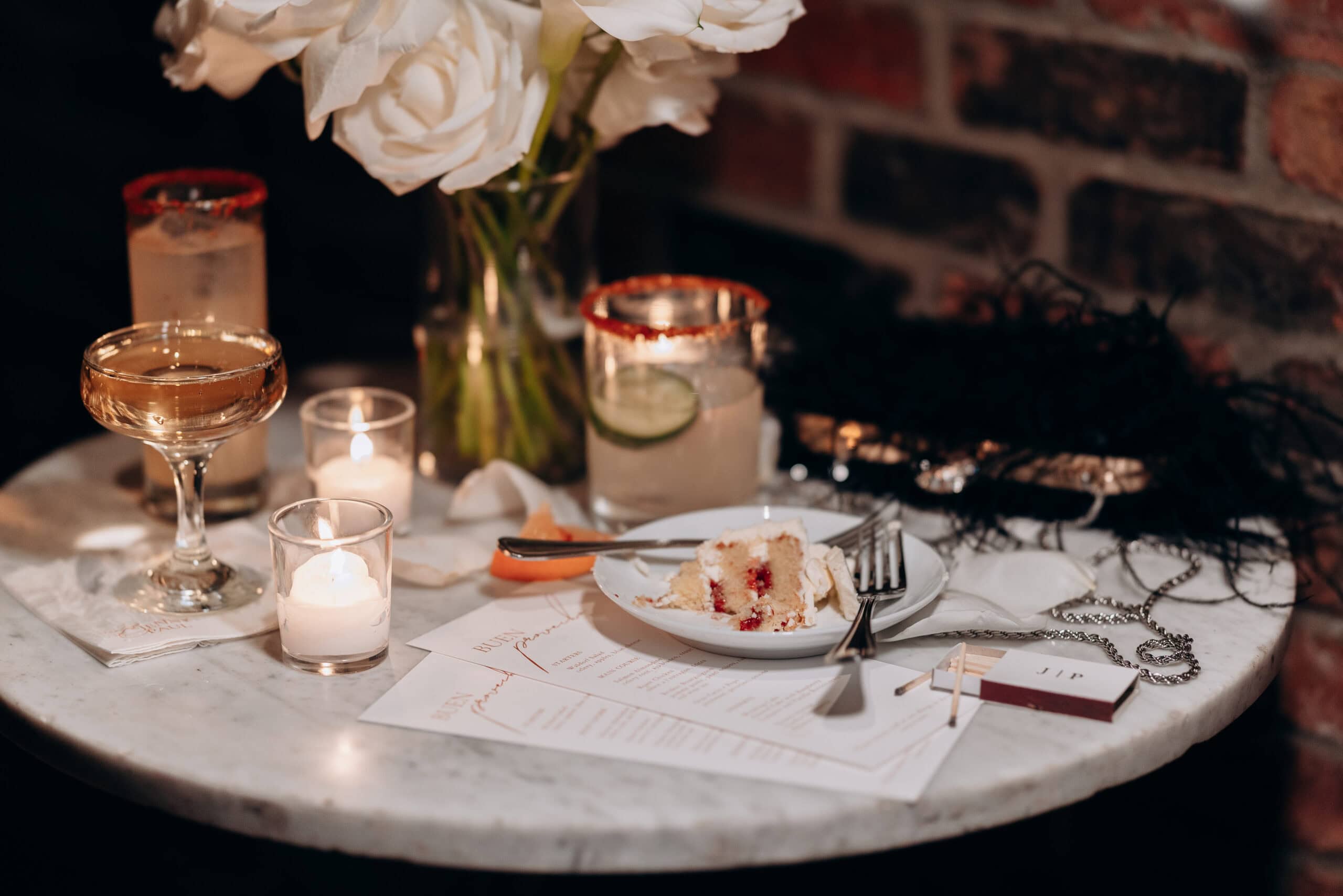 Close-up of a marble table with white roses in a glass vase, lit candles, a slice of layered cake on a white plate, cocktail glasses, and a brick wall background at Wythe Hotel, Brooklyn.