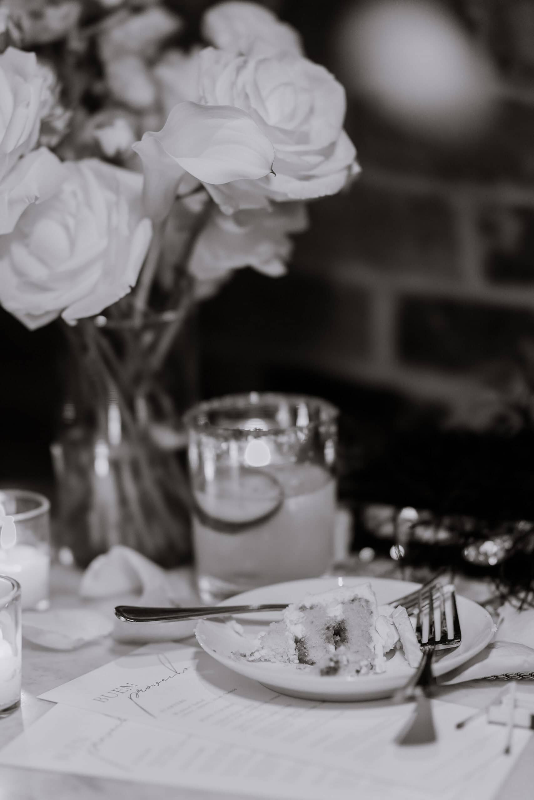 Close-up of white roses and calla lilies in a glass vase beside lit candles and a plate with partially eaten layered cake on a table at a wedding reception.