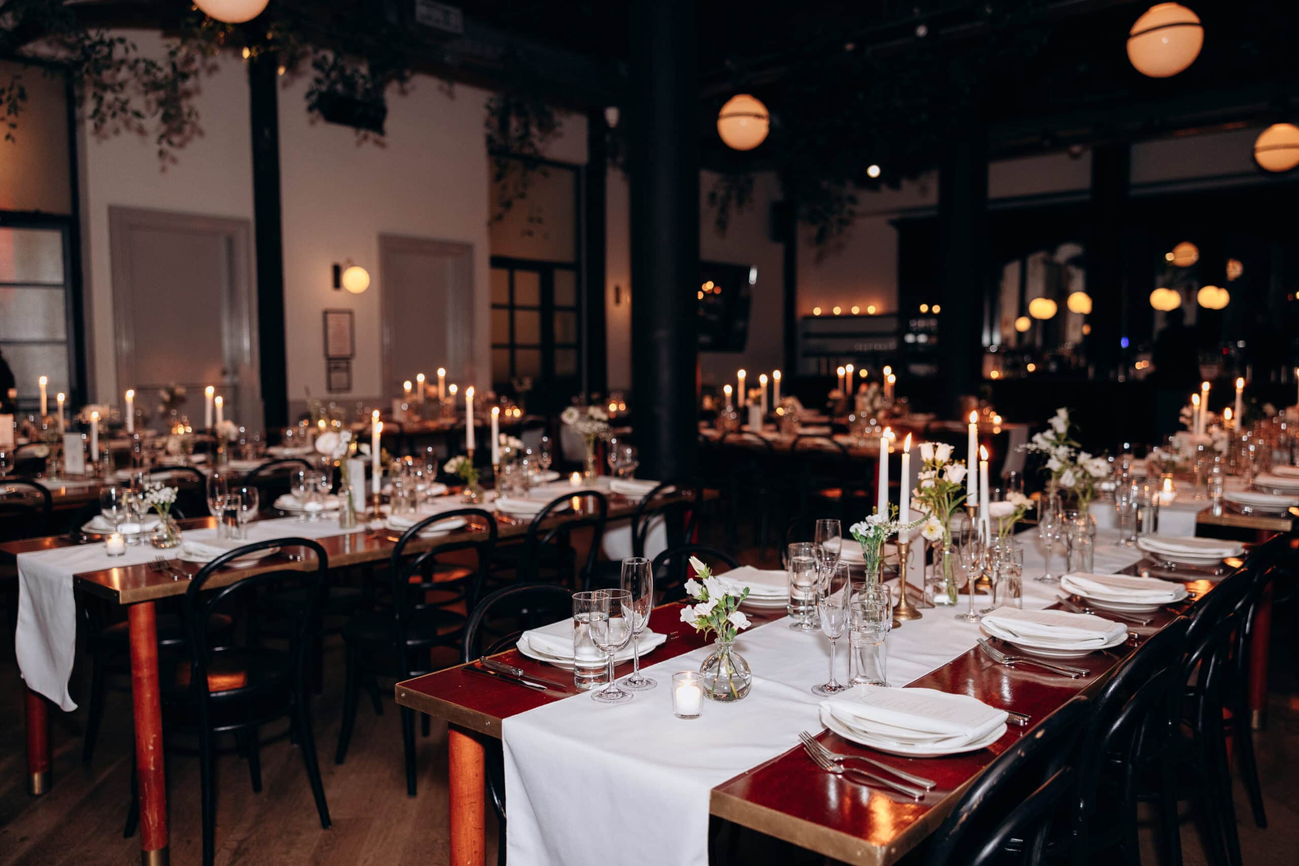 Candlelit wedding reception tables set with white linens, glassware, and floral arrangements in a softly lit Wythe Hotel dining room, Brooklyn