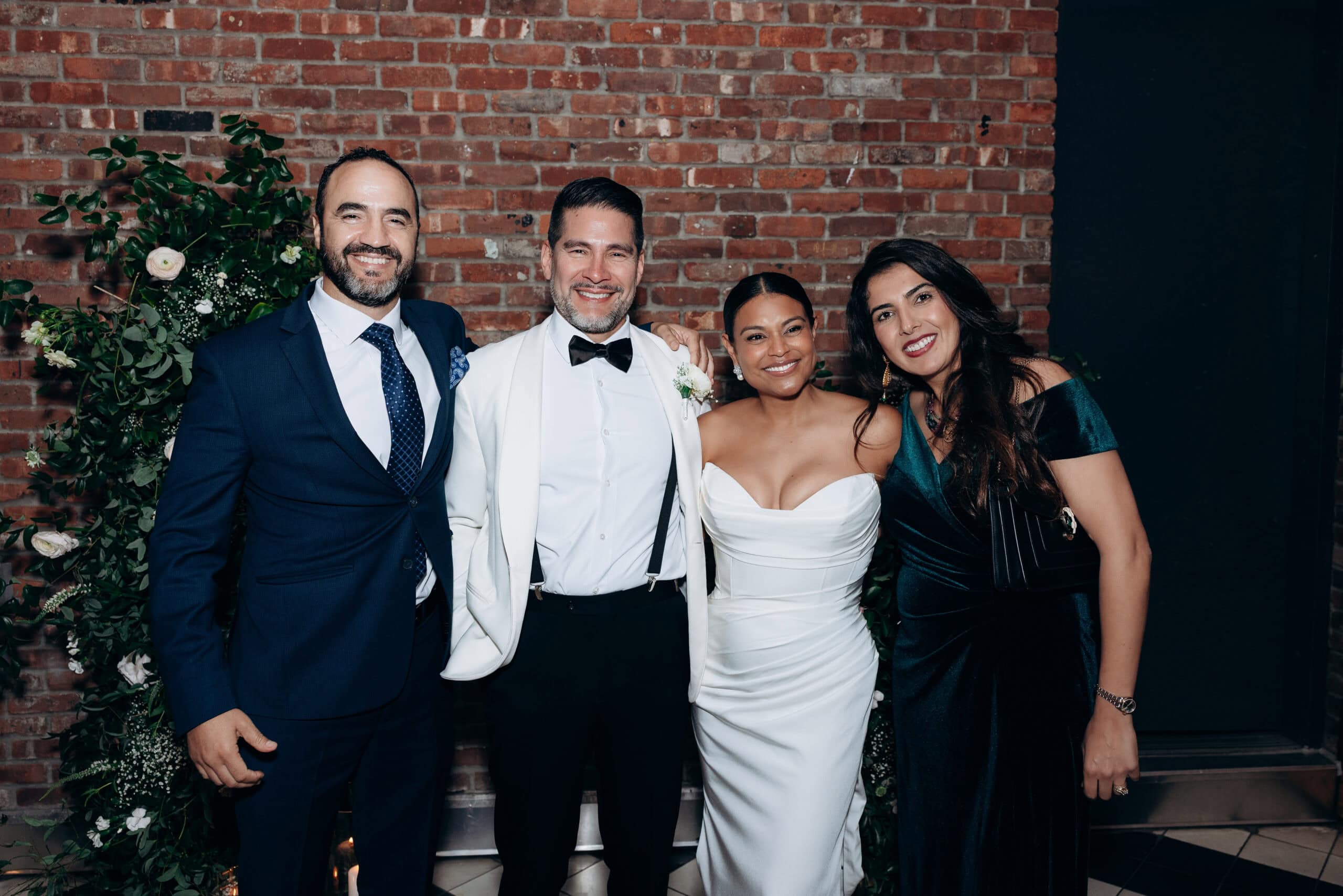 Four people posing in front of a brick wall with greenery; groom in white tuxedo jacket, bride in white gown, two guests in dark formal attire.