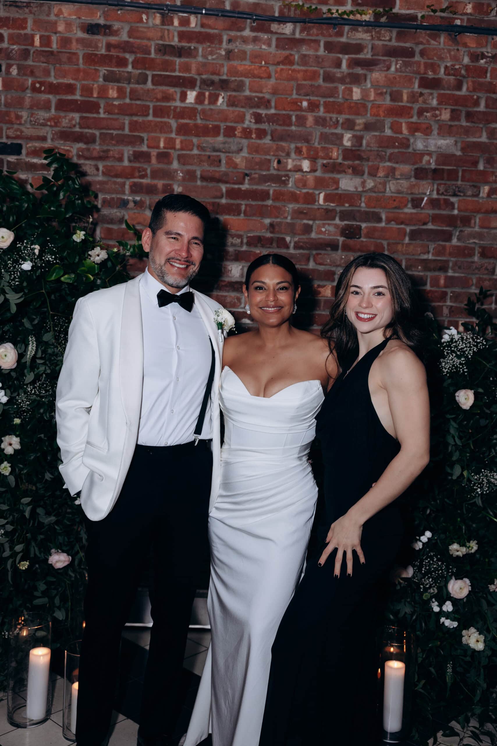 Three people posing in front of a red brick wall with floral greenery and lit candles; man in white tuxedo jacket, woman in white strapless gown, woman in black sleeveless dress.