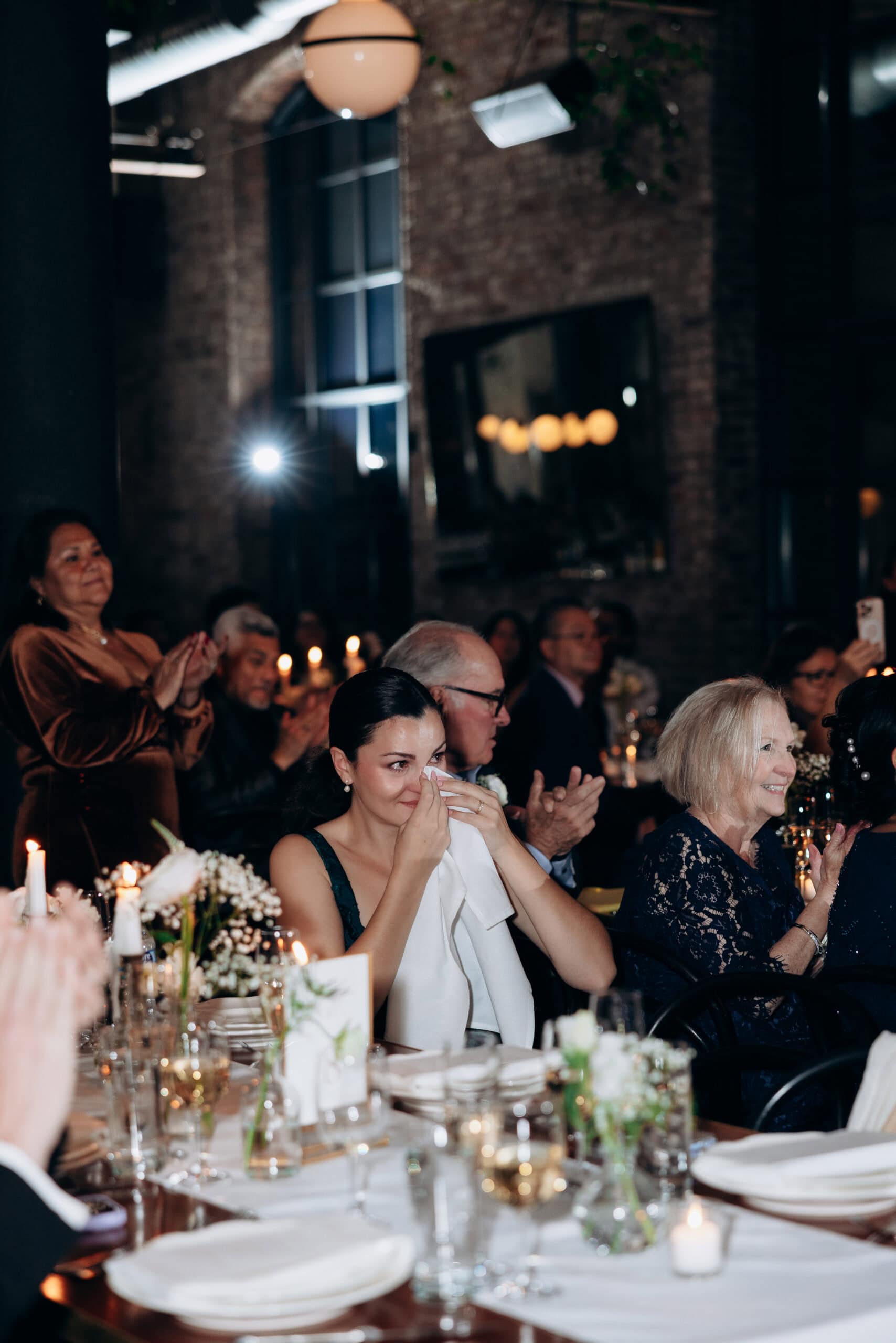 Woman wiping tears with a napkin during an emotional moment at a candlelit wedding reception in an industrial brick venue.