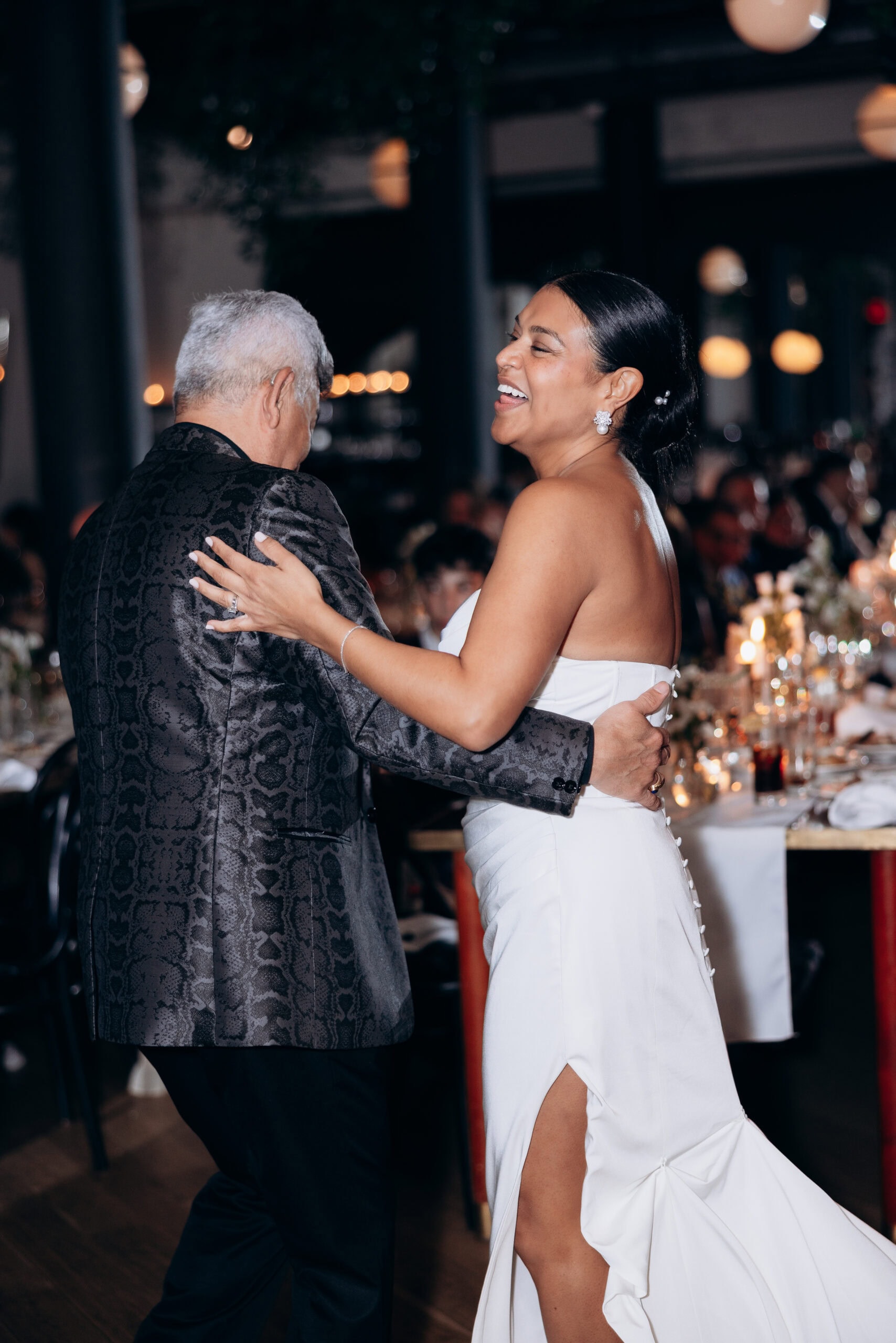 Bride in a white strapless gown with pearl earrings dancing closely with an older man in a dark patterned jacket at a dimly lit wedding reception.