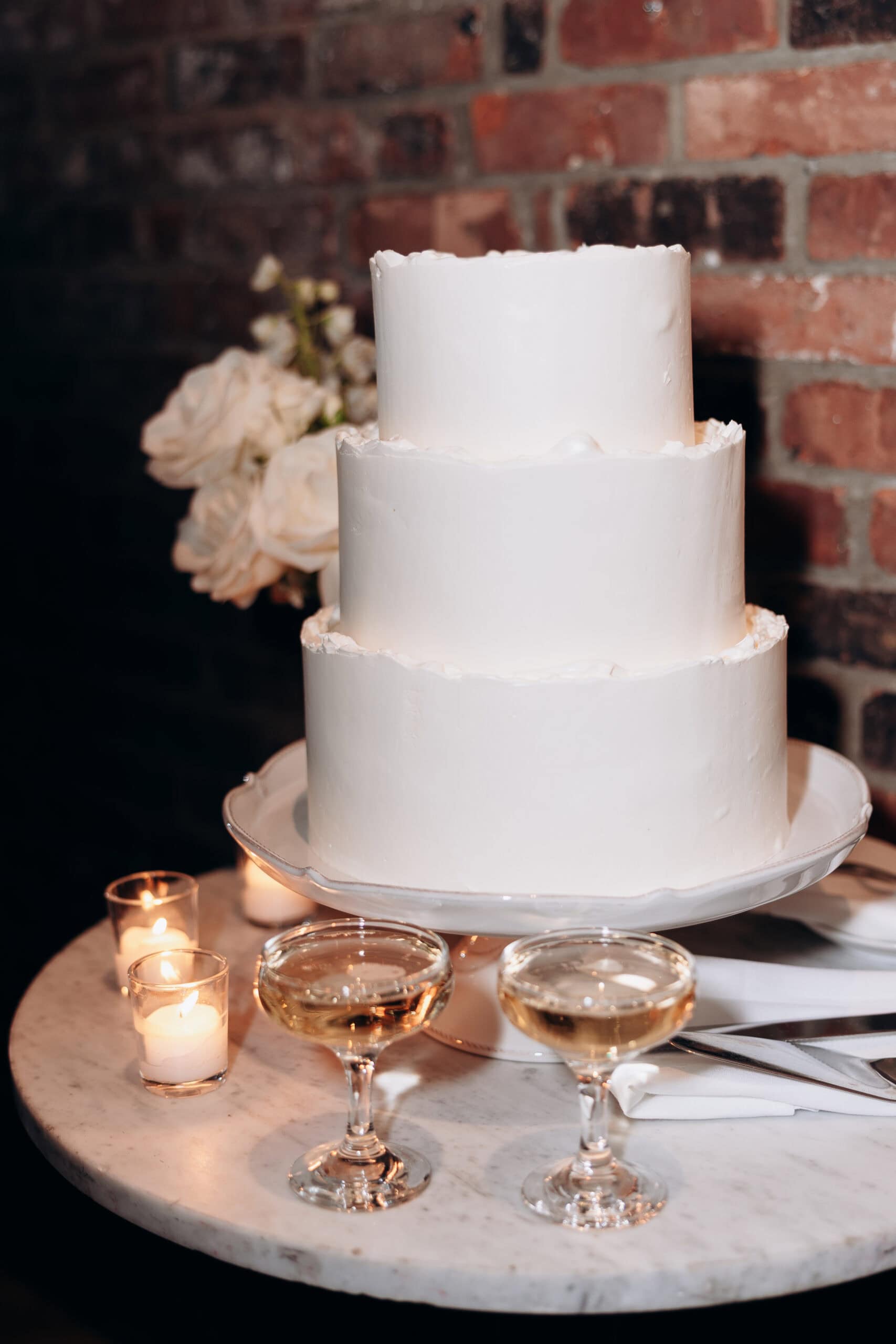 Three-tier white wedding cake on marble table with two champagne glasses, white roses, and lit votive candles in front of a brick wall at Wythe Hotel, Brooklyn.