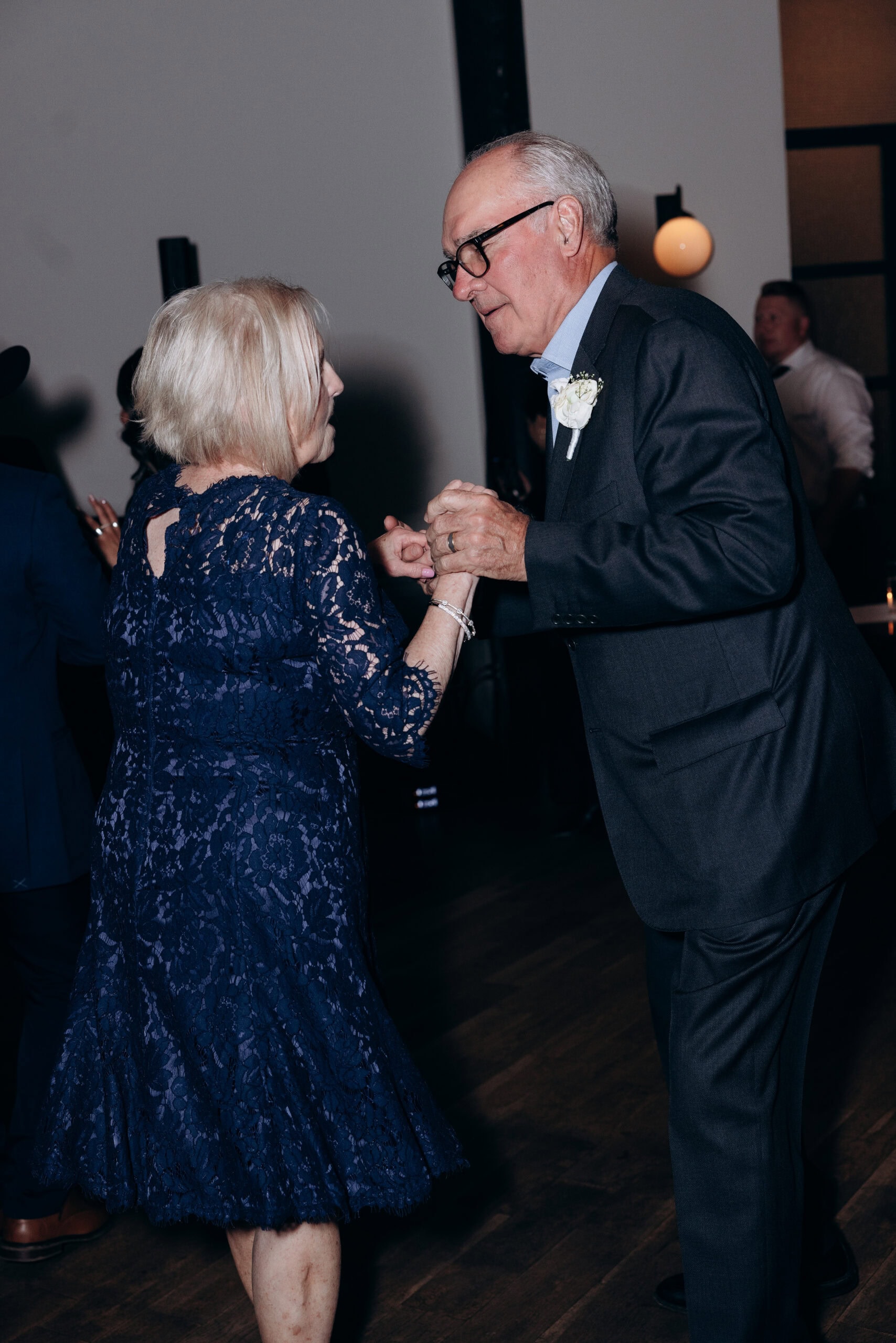 Older couple dancing closely indoors, woman in navy lace dress and man in dark suit with boutonniere, dimly lit venue with warm wall sconce.