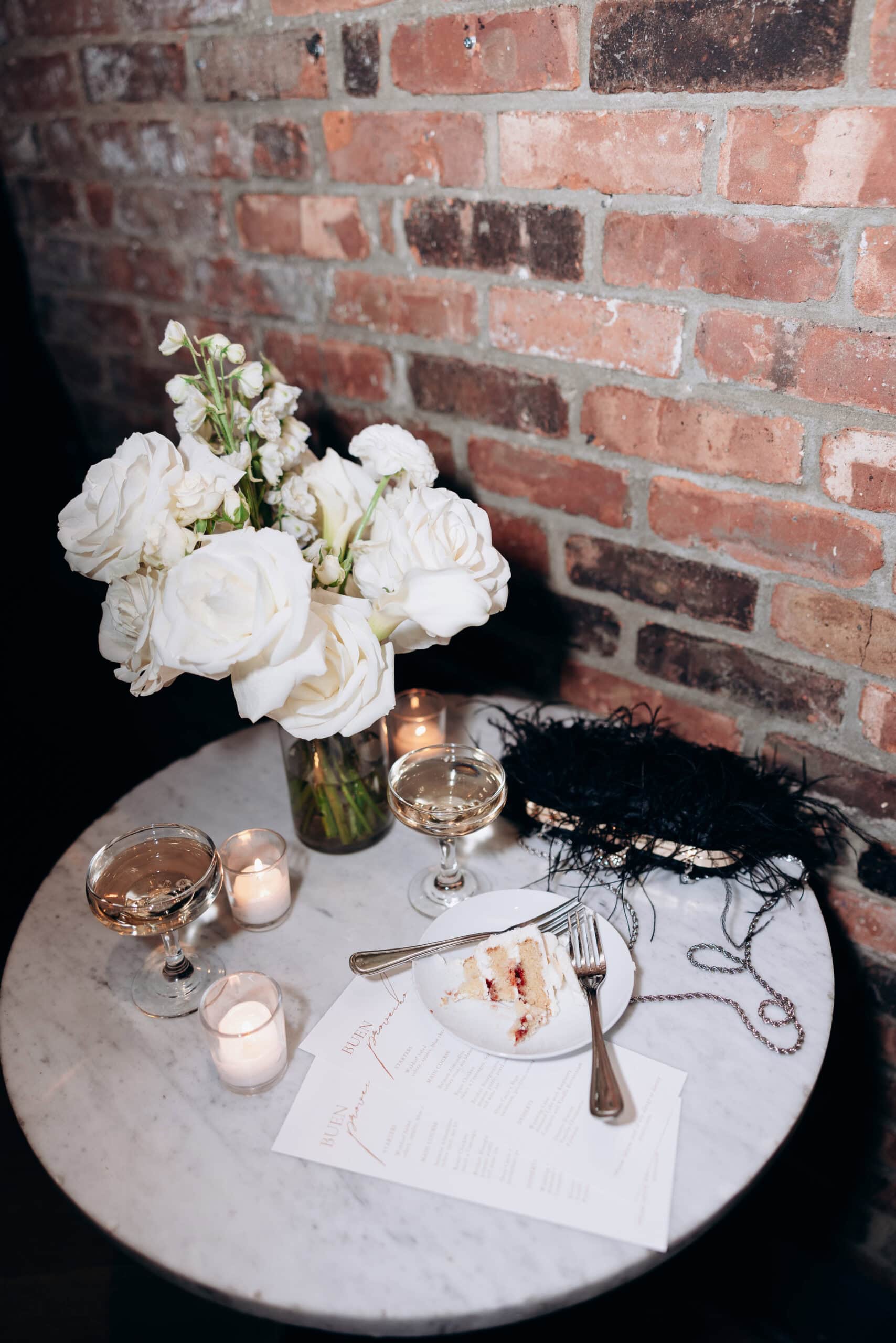 White floral bouquet in glass vase, slice of layered cake with fork on white plate, two coupe glasses with champagne, votive candles on marble table against exposed brick wall.