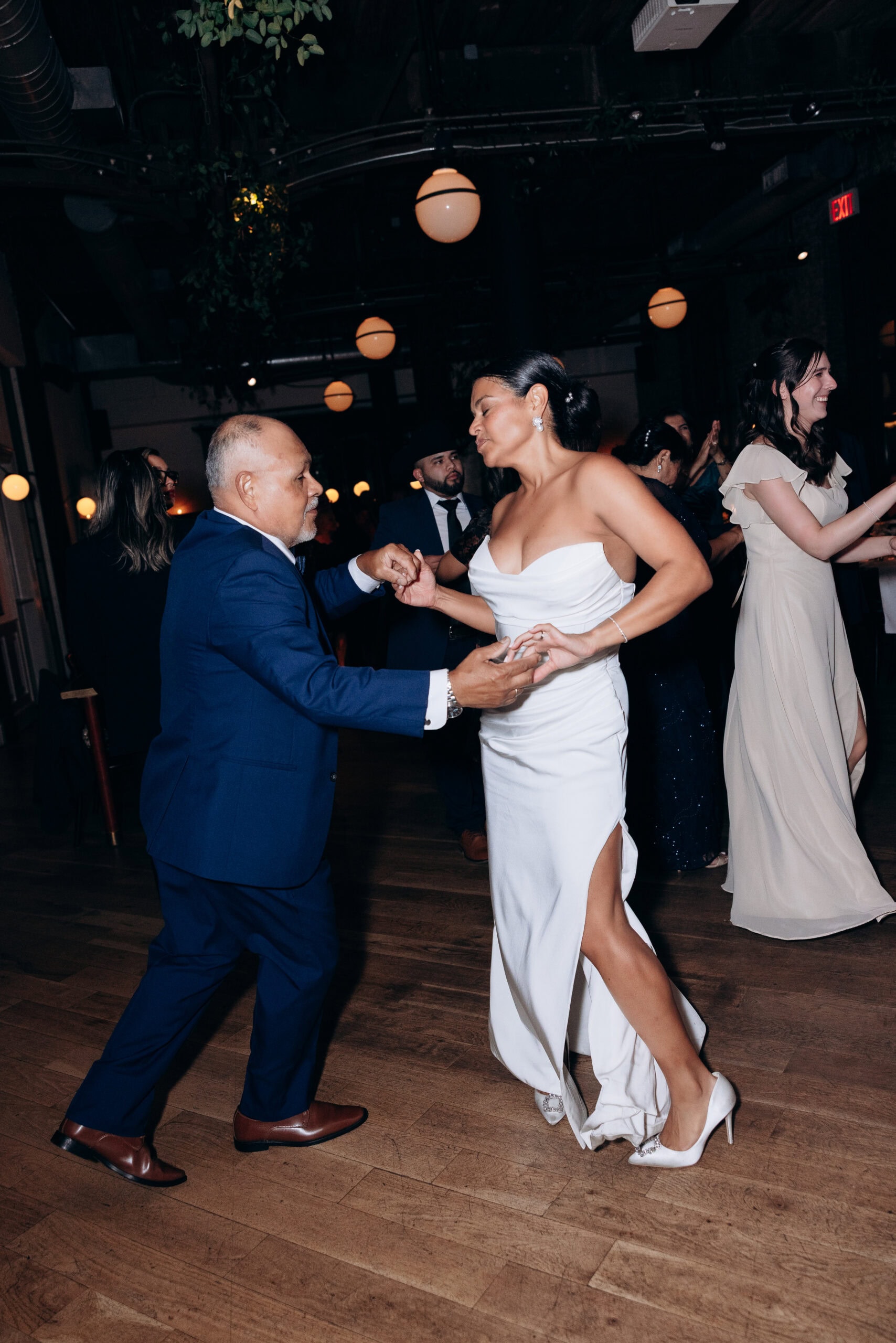 Bride in white strapless gown dancing with older man in navy suit under warm round lights at Wythe Hotel, Brooklyn wedding reception.