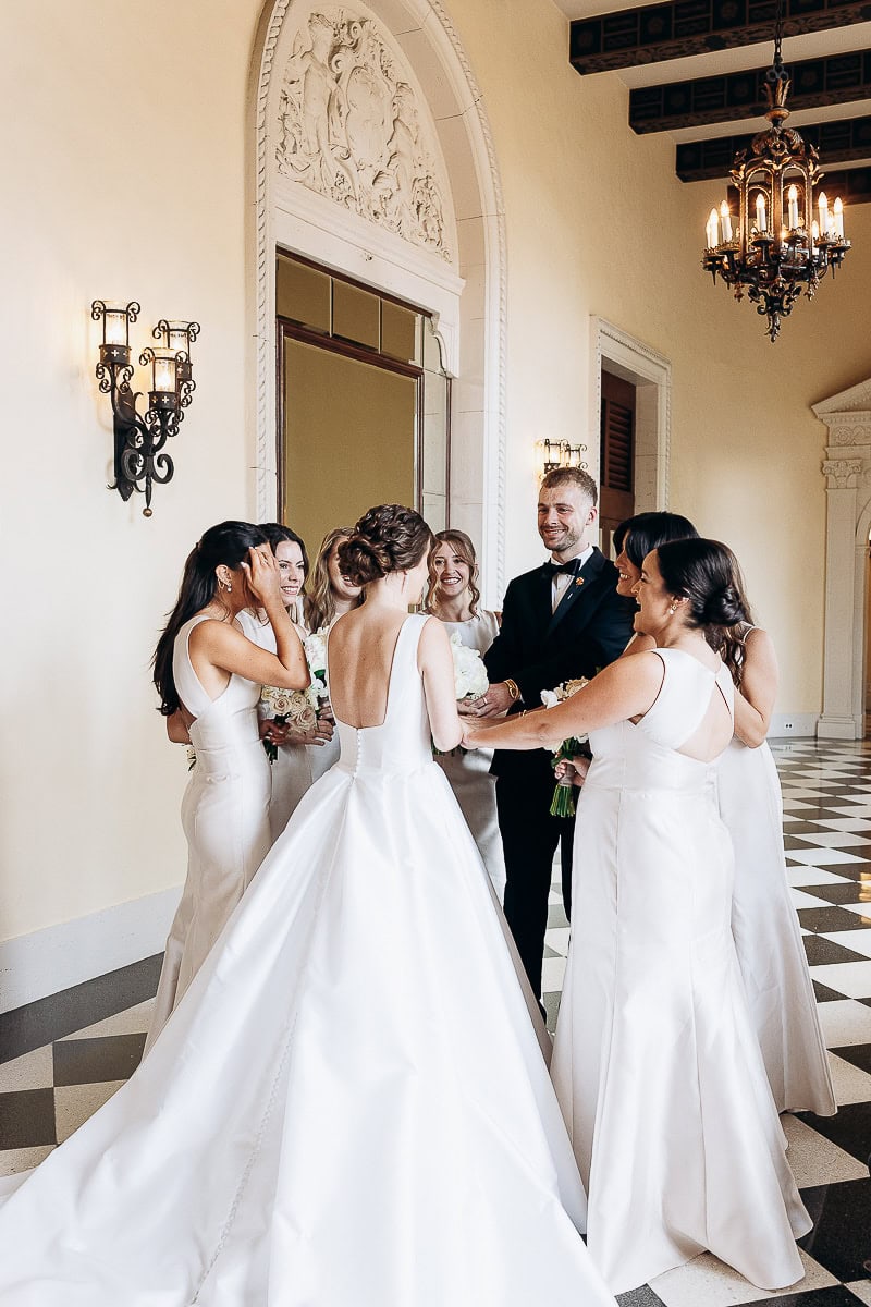 Bride in a satin gown surrounded by bridesmaids and a groomsman in a grand hall with ornate wall sconces and chandelier at Westchester Country Club, Rye, NY.