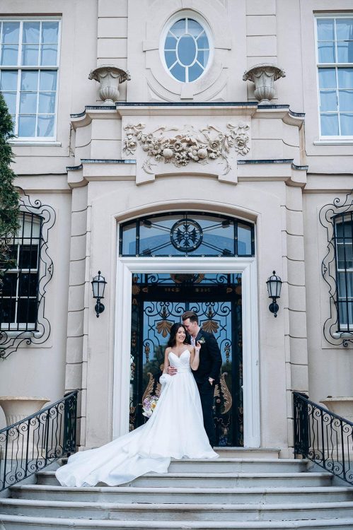 Bride and groom embracing on steps of a grand Manhattan mansion with intricate wrought iron doors and classical stone facade.