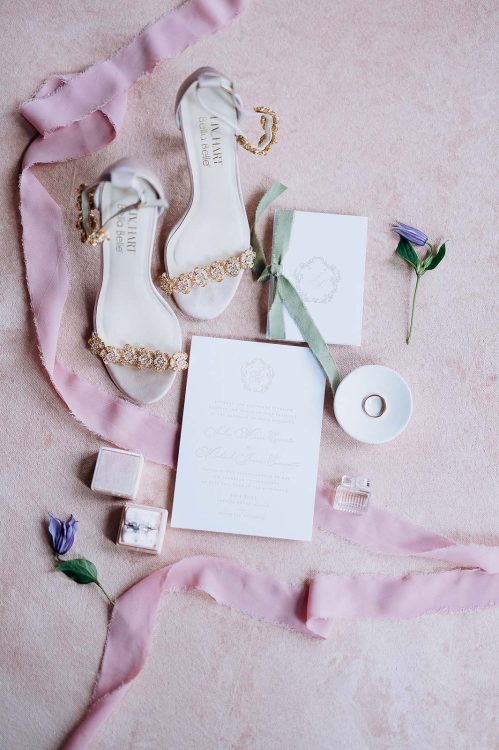 Flat lay of luxury wedding details including embellished ivory heels, invitation suite with sage ribbon, wedding ring on white dish, velvet ring box, and mauve silk ribbons on blush fabric.
