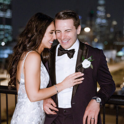 Bride in floral lace gown and groom in black tuxedo with bow tie, sharing a joyful moment at night with Manhattan skyline blurred in background.