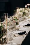 Close-up of a wedding table setting with lit taper candles in glass holders, crystal wine glasses, white plates with black napkins, and soft floral arrangements at Westchester Country Club, Rye, NY.
