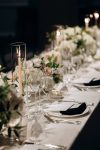 Close-up of a wedding table setting with lit taper candles in glass holders, crystal wine glasses, white plates with black napkins, and soft floral arrangements at Westchester Country Club, Rye, NY.