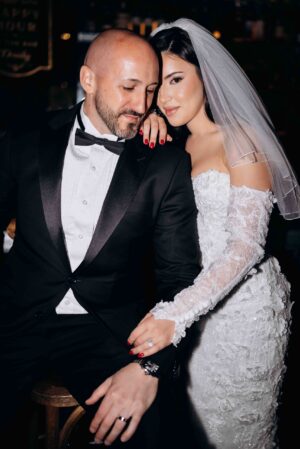 Bride in lace wedding gown and veil gently resting her hand on groom's shoulder, groom in black tuxedo with bow tie, seated on wooden stool in dimly lit setting, New York, NY.