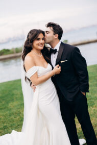 Bride in off-shoulder white gown and pearl necklace embraces groom in black tuxedo as he kisses her cheek near waterfront in New York, NY.
