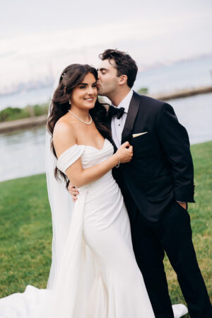 Bride in off-shoulder white gown and pearl necklace embraces groom in black tuxedo as he kisses her cheek near waterfront in New York, NY.